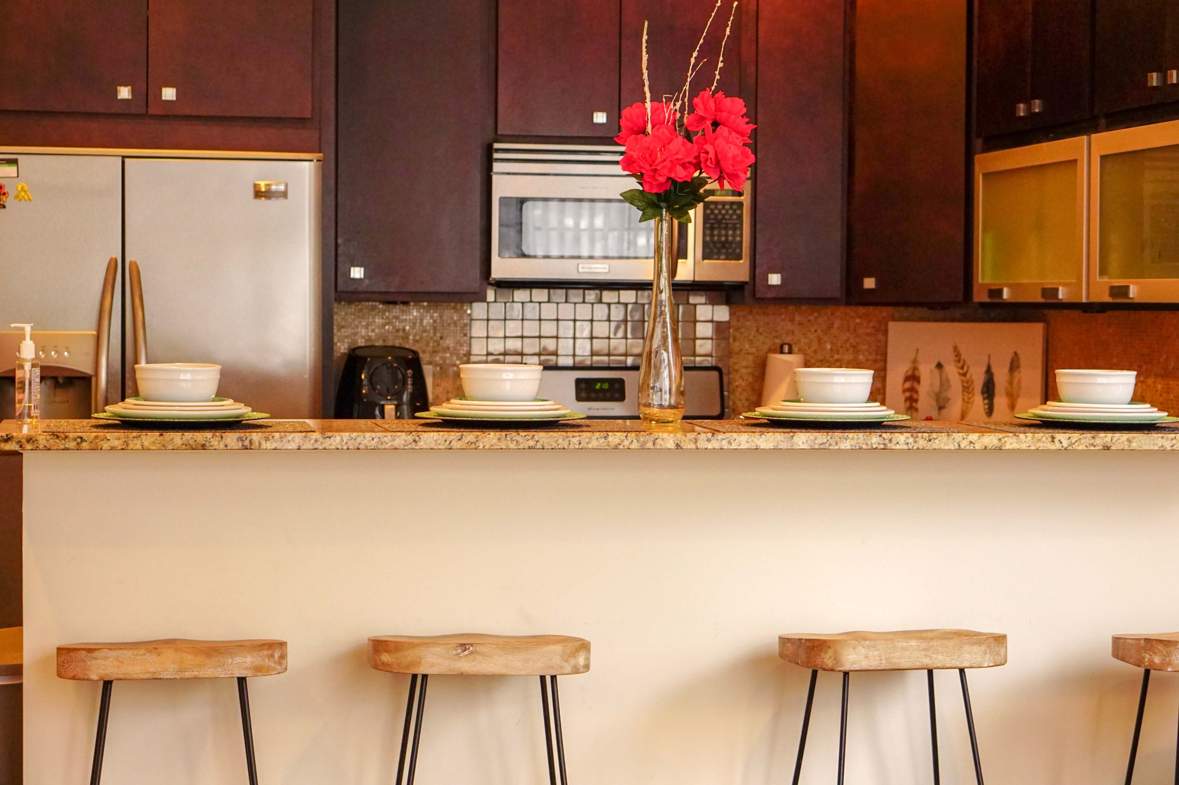 A kitchen bar area features four wooden stools, a granite countertop, and a vase of flowers, with plates and bowls set for a meal.