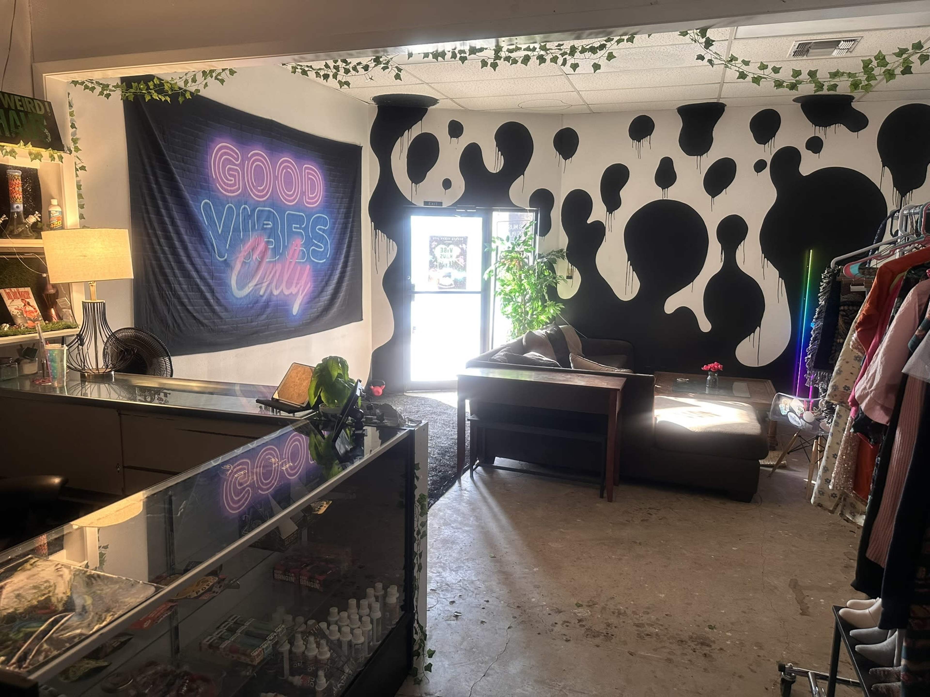 The image shows a shop interior featuring a colorful "Good Vibes Only" banner on a wall with black and white abstract patterns, a couch, a plant, and a clothing rack.