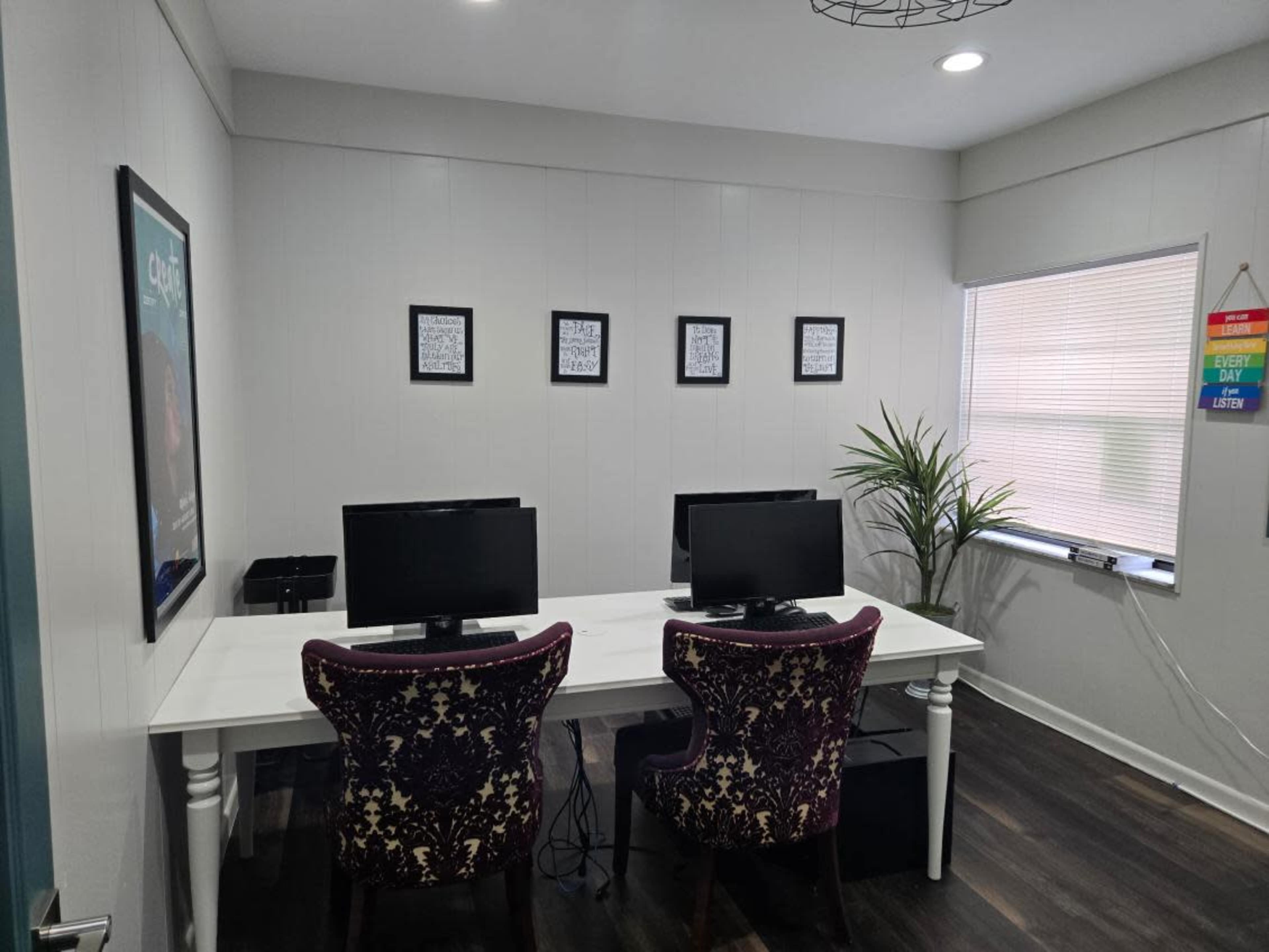 The image shows a modern office space with two computer workstations facing a white table, adorned with several framed artworks and a potted plant on one side.
