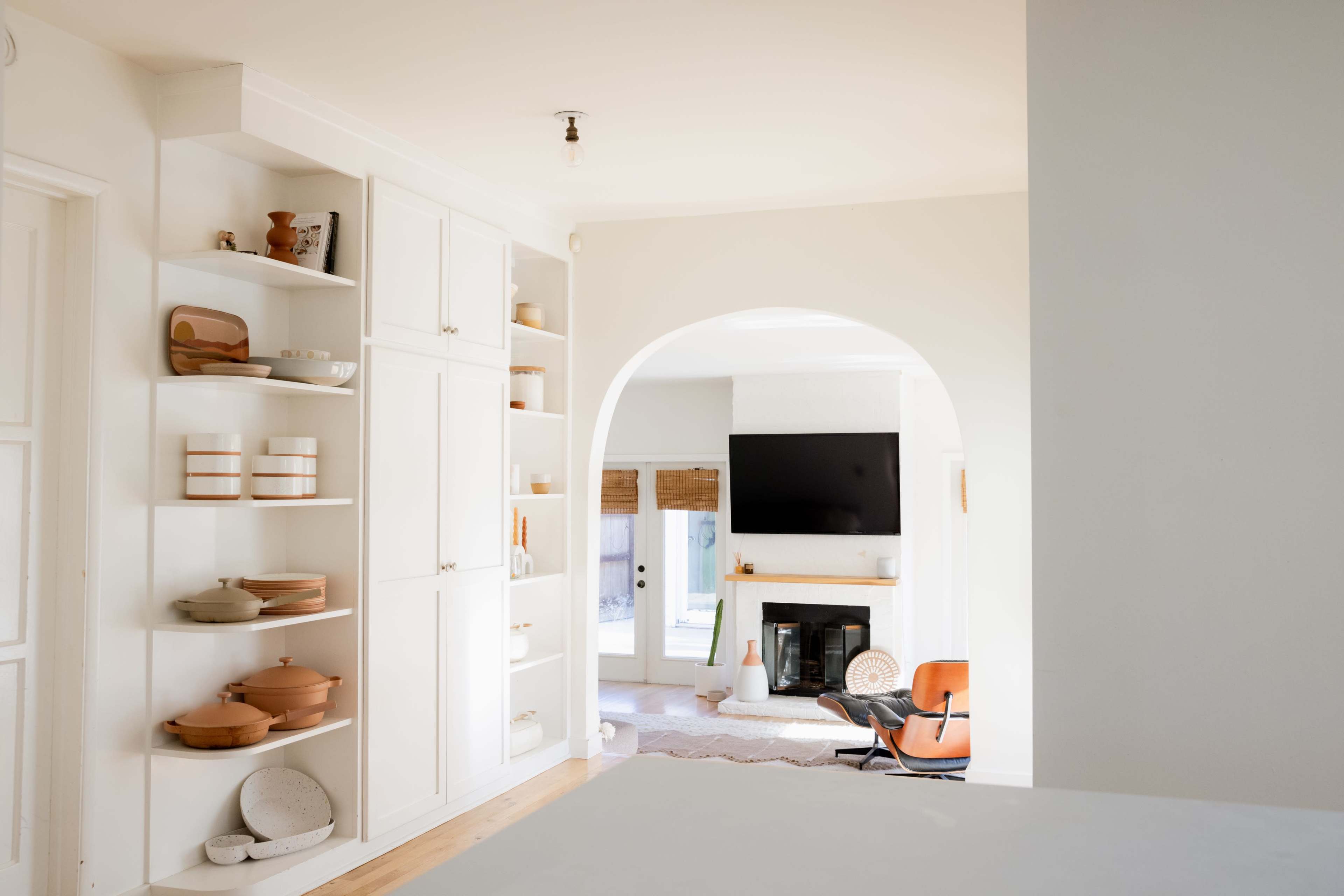 A bright, airy living space features built-in shelves with dishware, leading to a cozy sitting area with a fireplace and a large television.