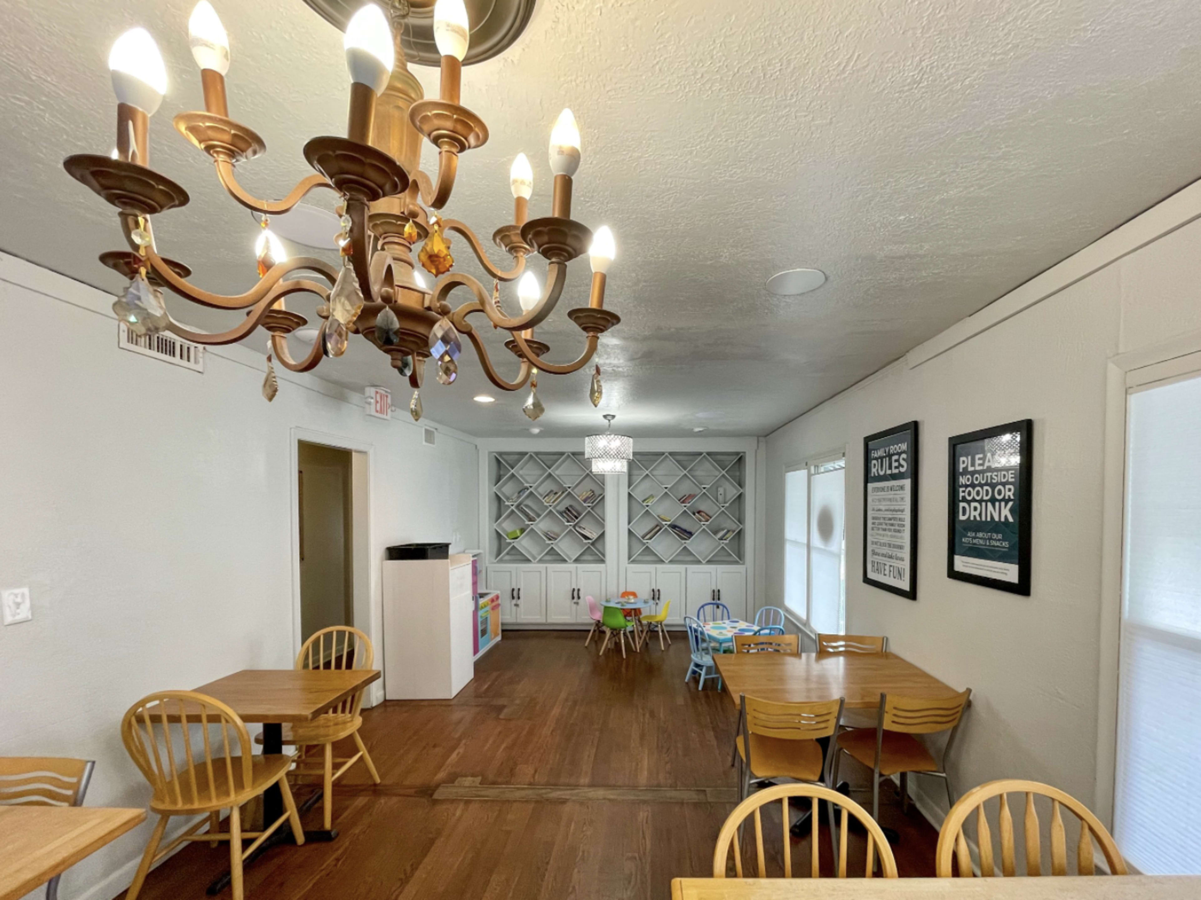 The image shows a dining area with wooden tables and chairs, a chandelier, and a backdrop featuring a white shelf and framed posters on the walls.