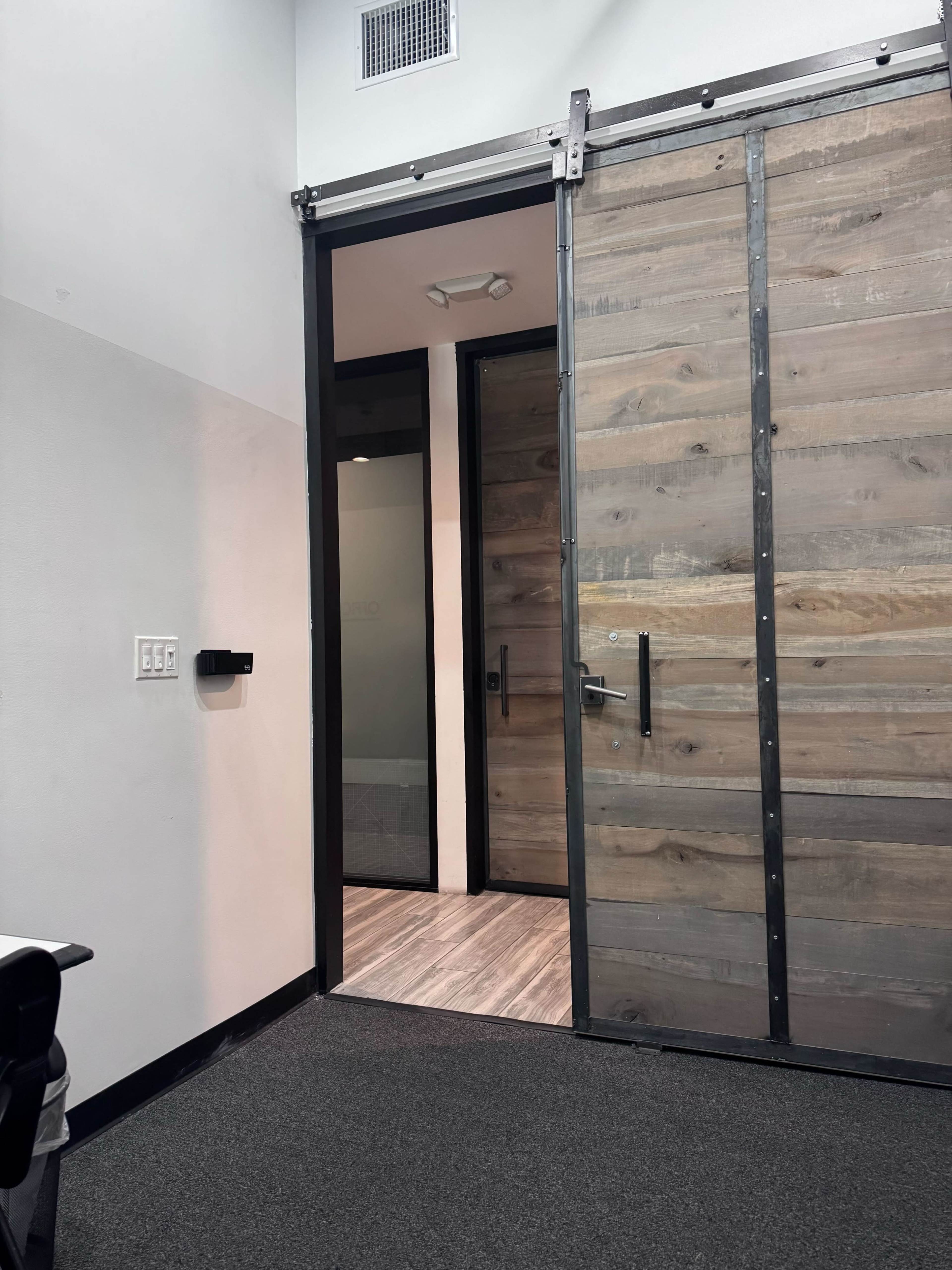 The image shows a partially open wooden sliding door revealing a hallway with gray flooring and a modern interior.