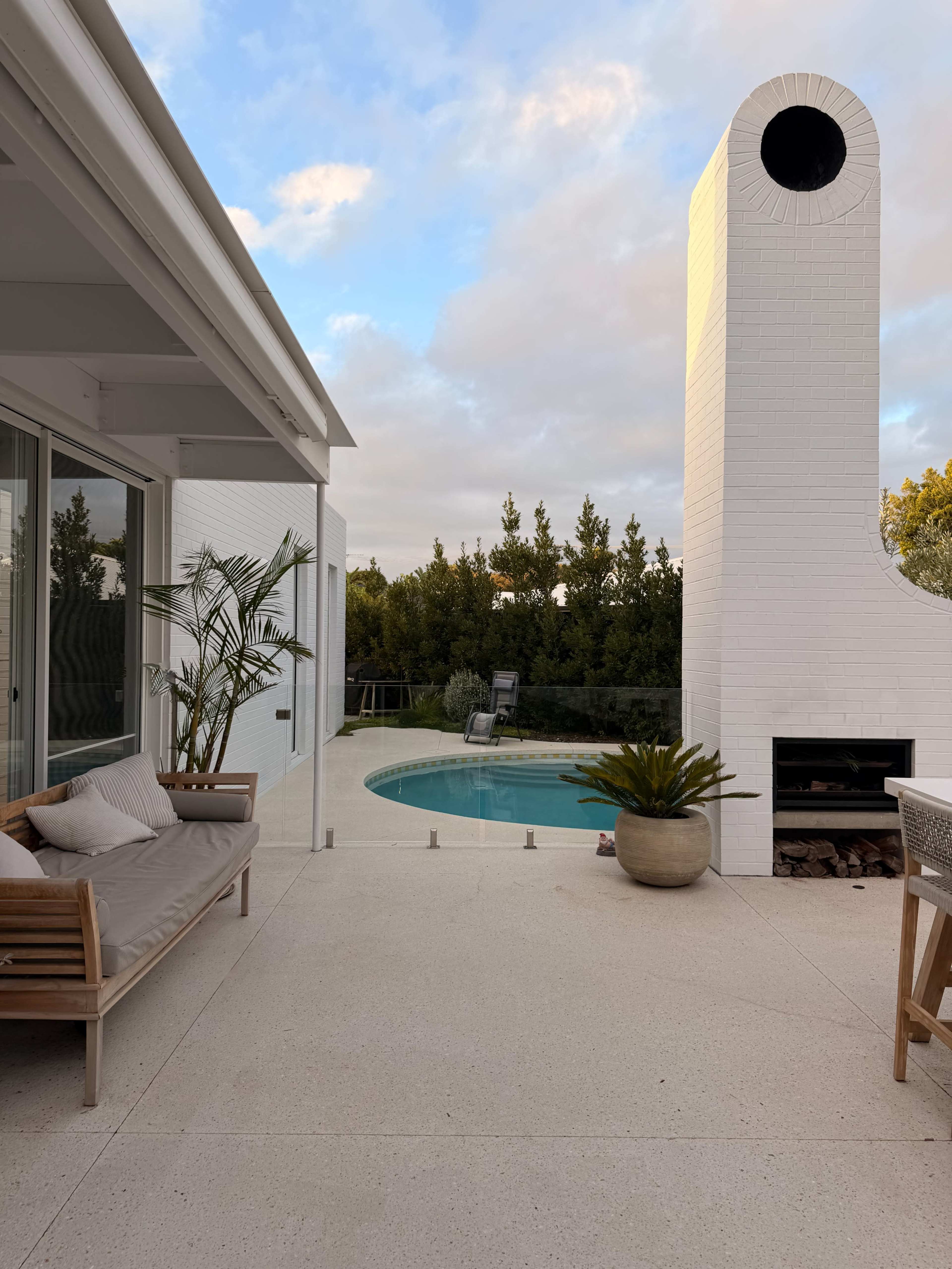 The image shows a modern outdoor patio area featuring a seating arrangement, a pool, and a tall white chimney.