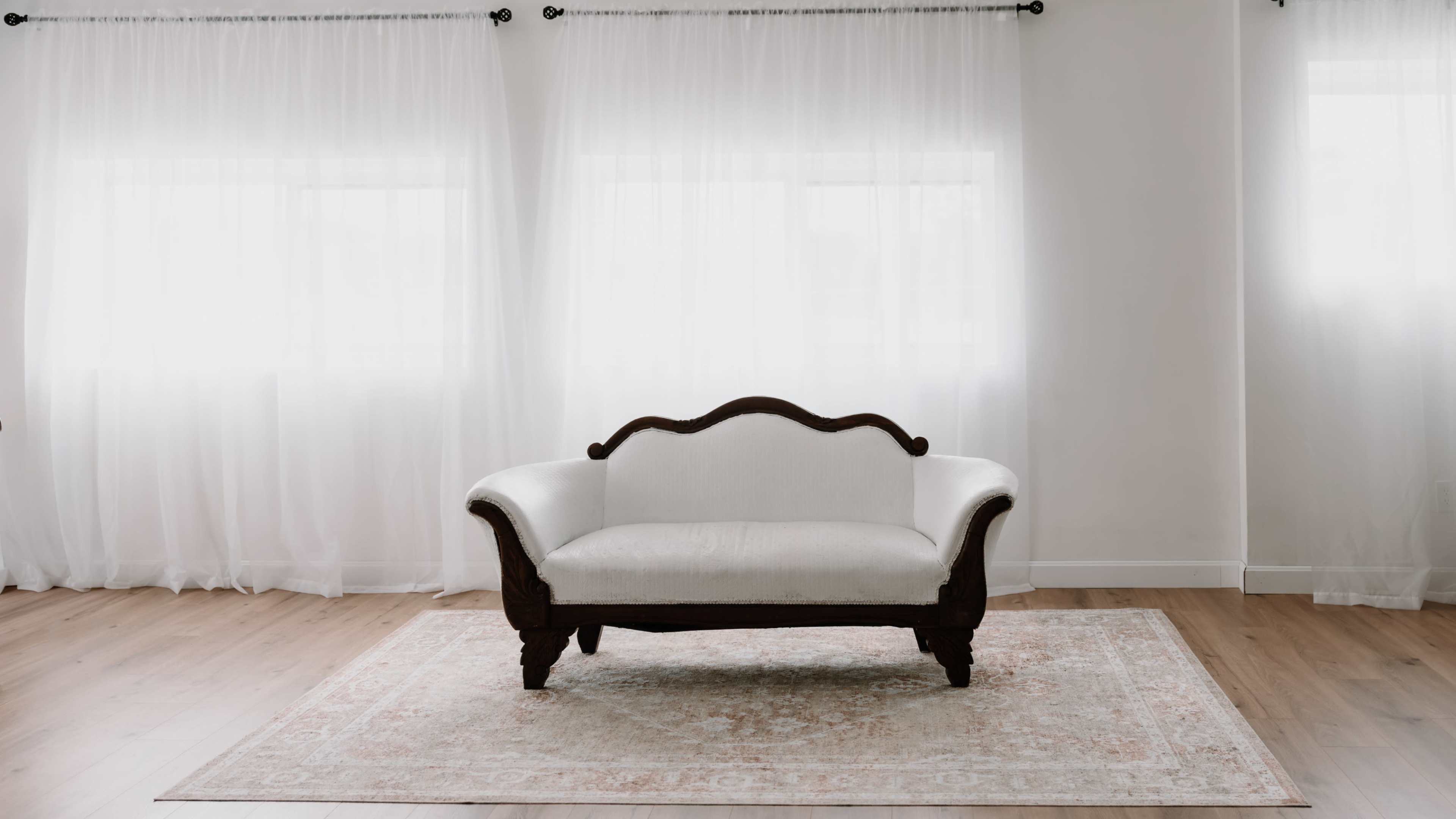 A white upholstered sofa with wooden legs is placed on a decorative rug in a room with sheer white curtains.