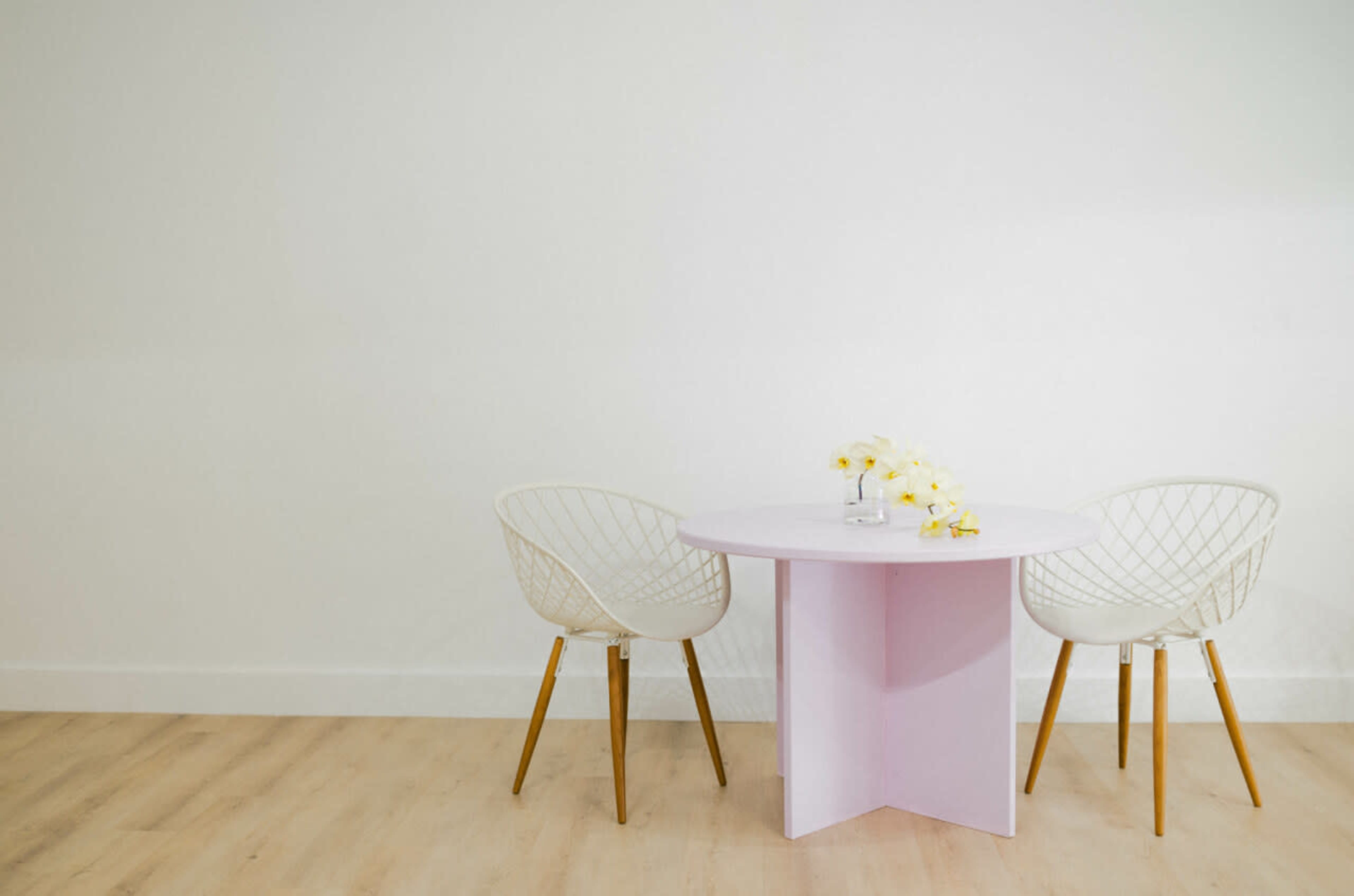 A round pink table with a vase of yellow flowers is set between two white wire chairs on a light wood floor against a plain white wall.