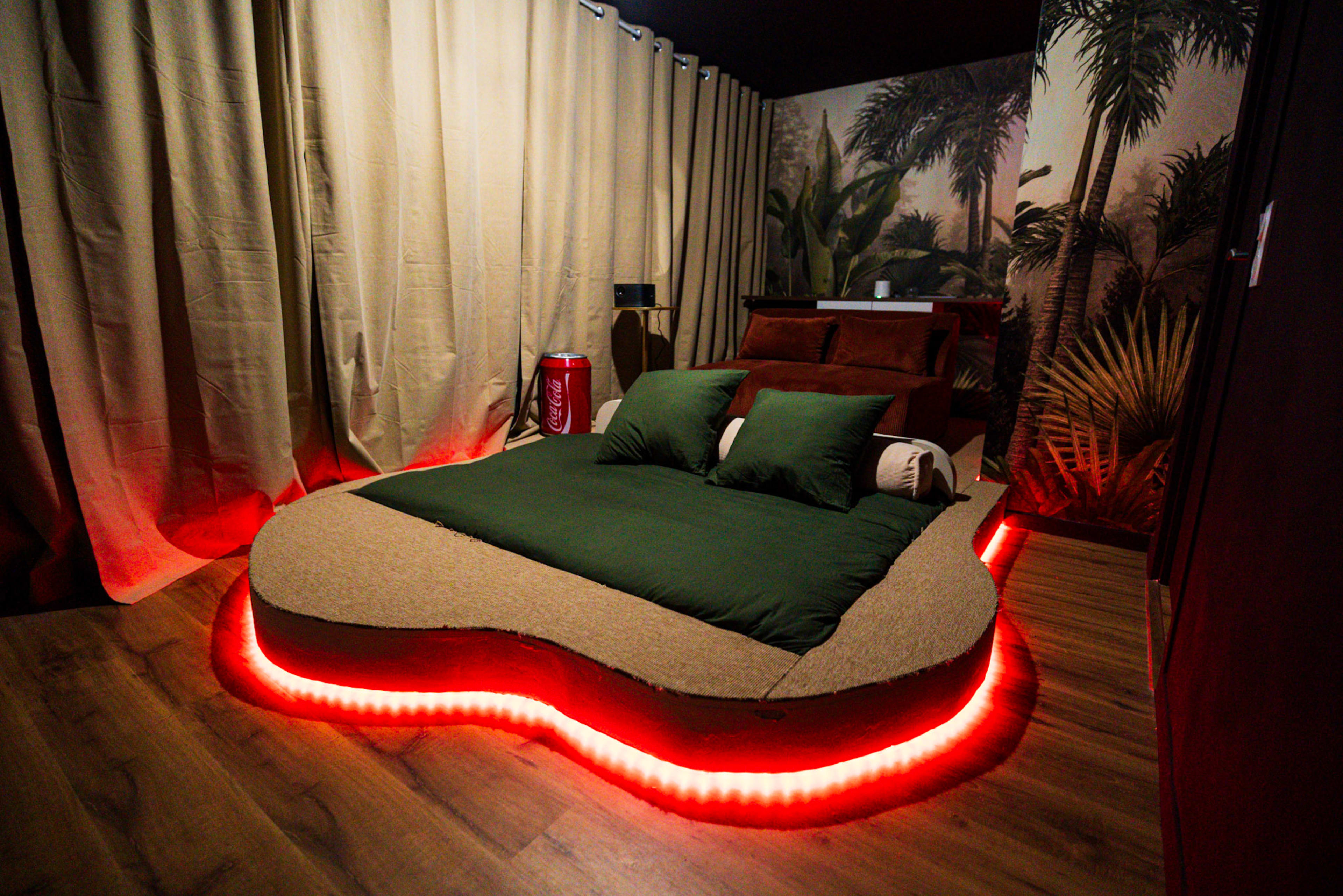 The room features a uniquely shaped bed with green bedding and a red LED light border, surrounded by beige curtains and a tropical-themed wall mural.