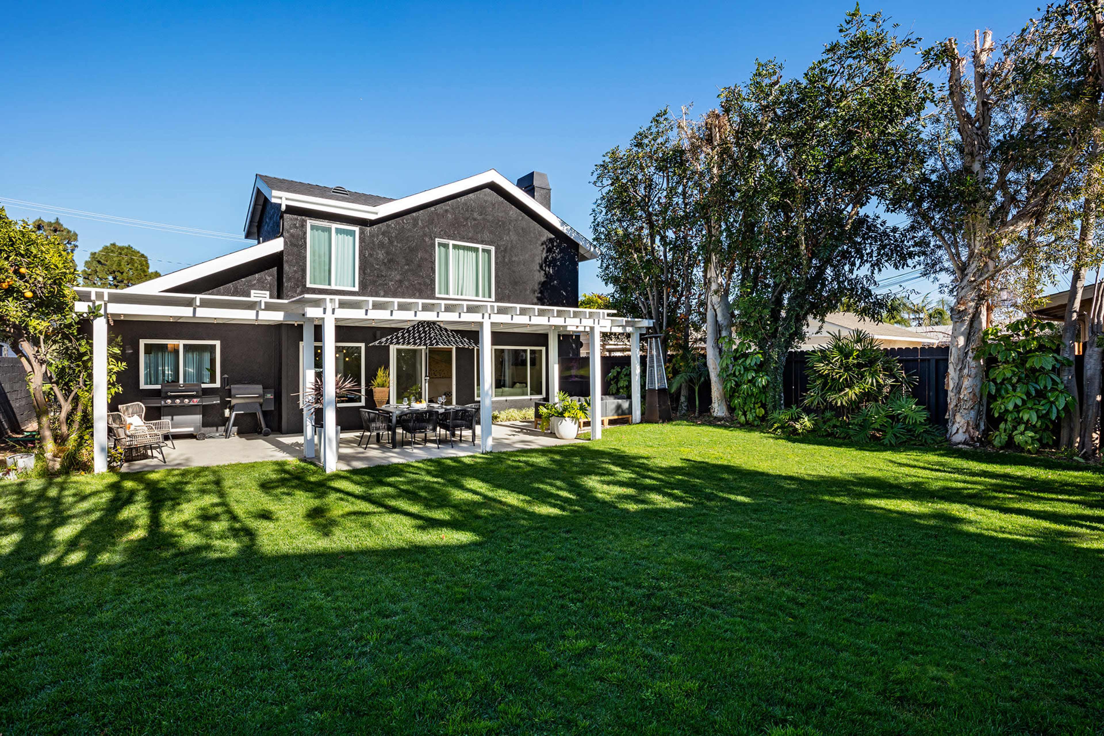 A two-story black house features a spacious backyard with a patio covered by a pergola and lush green grass.