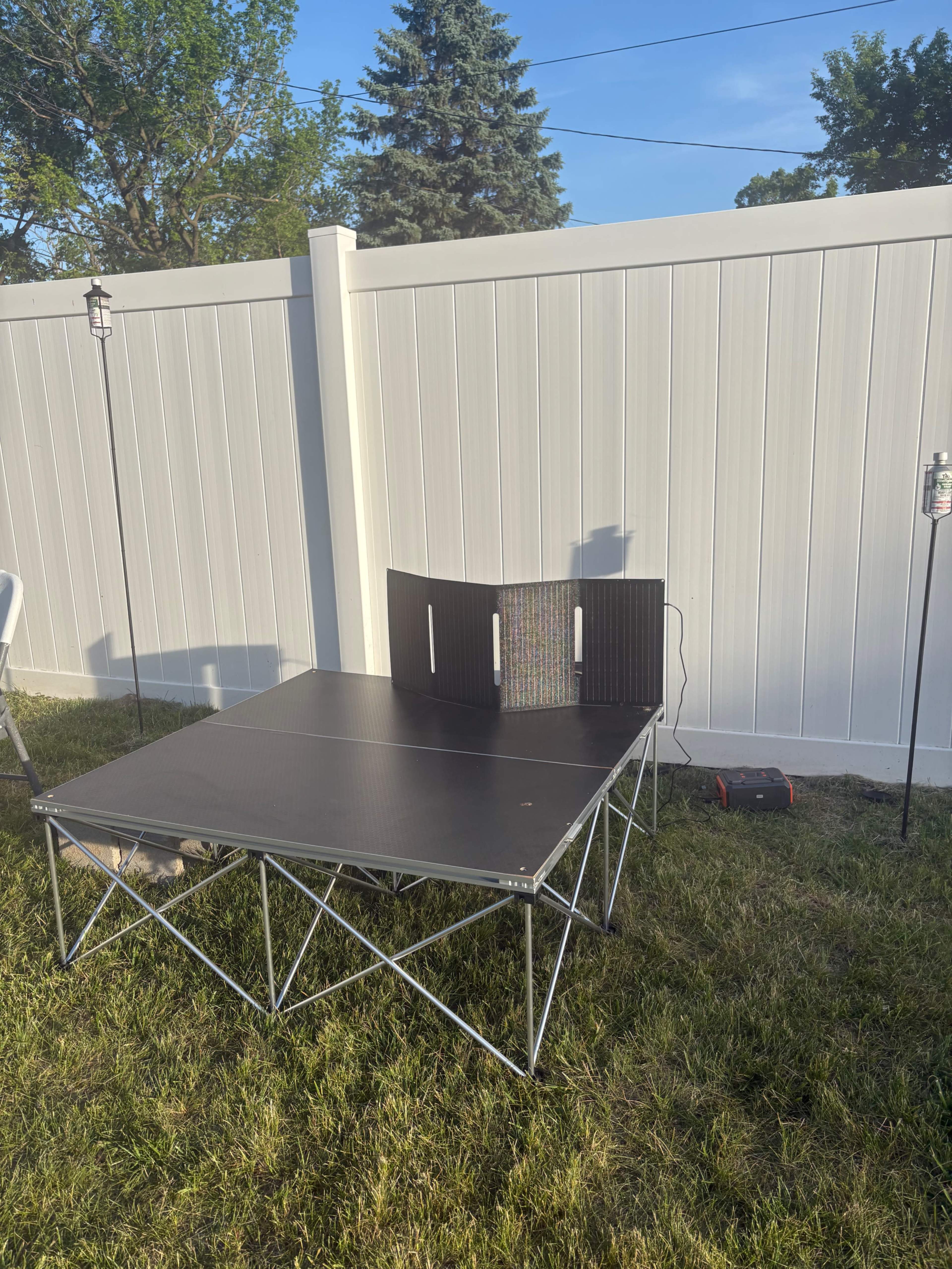 The image shows a portable black folding table set up on grass with a white vinyl fence in the background and two lamp posts nearby.