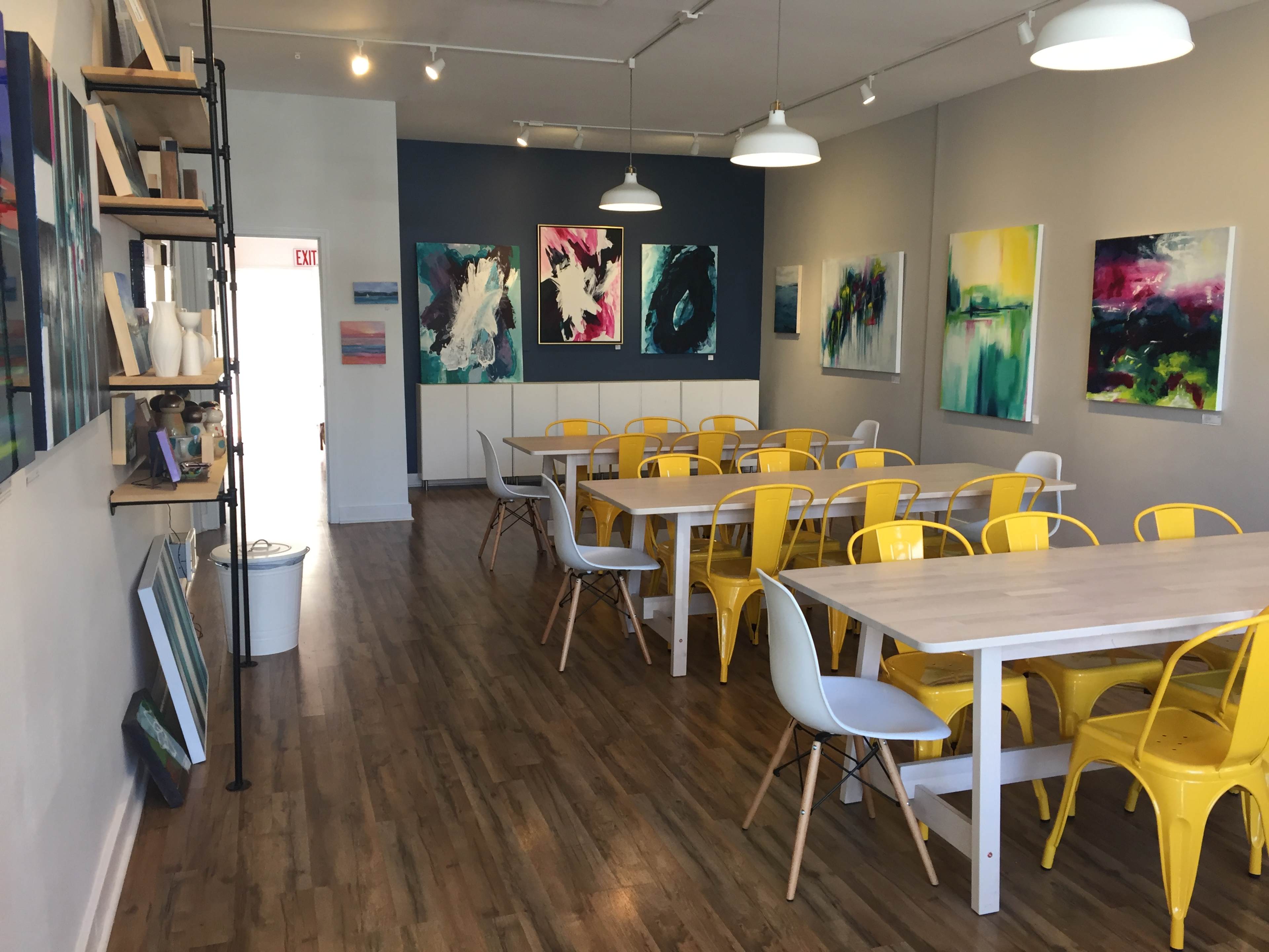 A bright, modern art studio with yellow chairs and wooden tables, adorned with colorful abstract paintings on the walls.