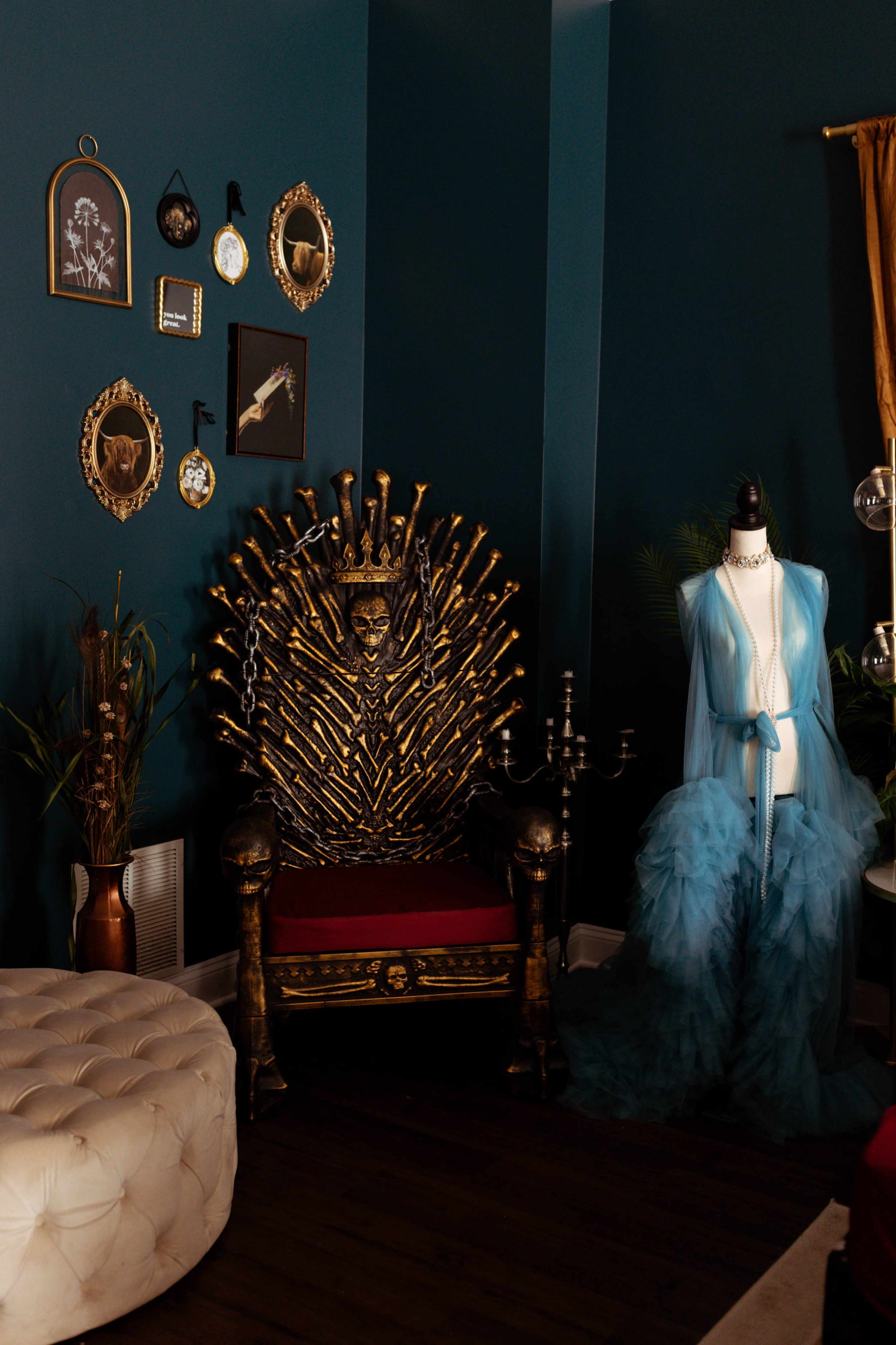 The image shows a decorative room featuring a throne made of swords, a mannequin dressed in a blue gown, and framed artwork on the dark teal wall.