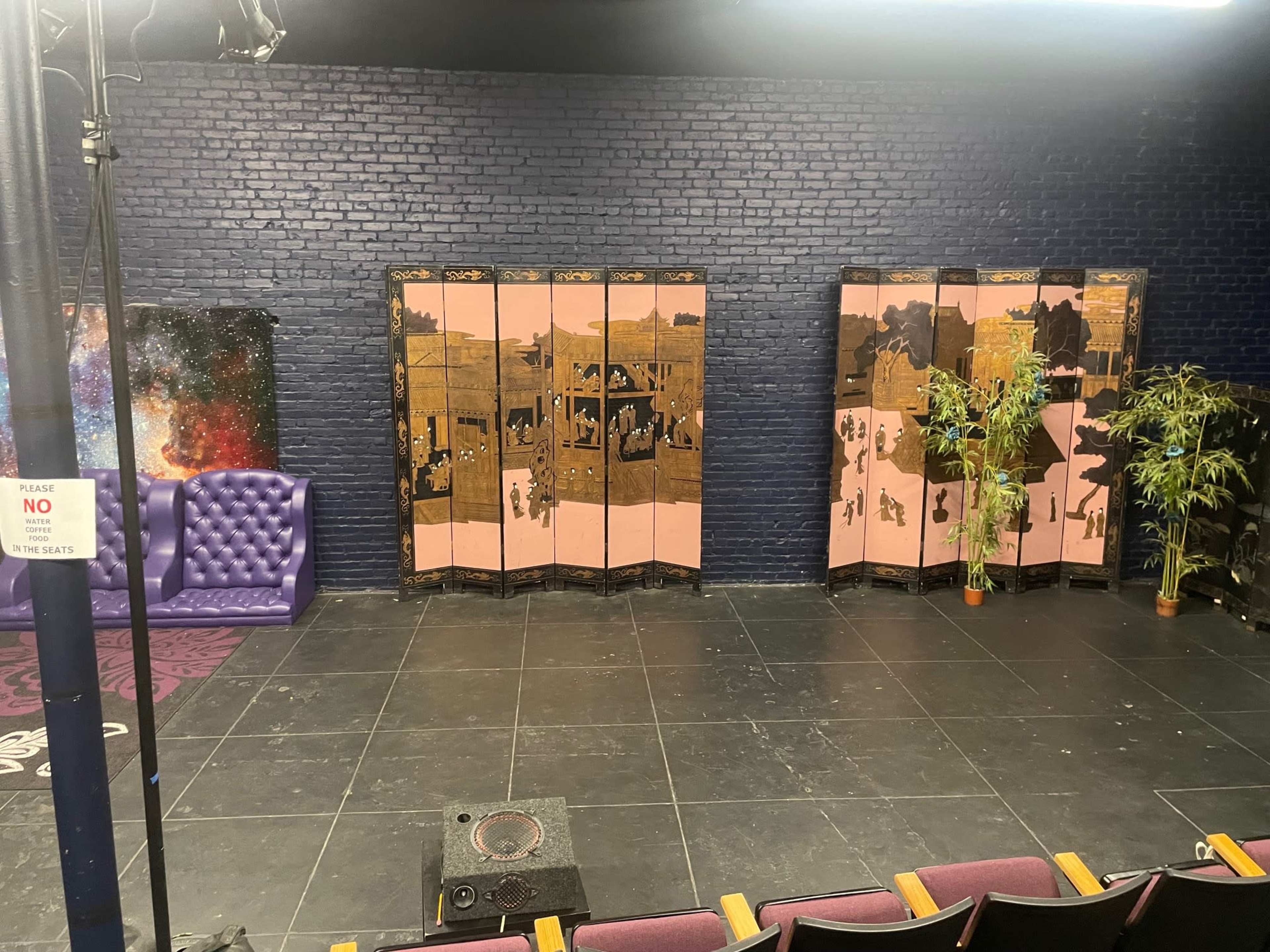 A stage area with a purple sofa, a black floor, folding screens featuring painted scenes, and potted plants on either side.