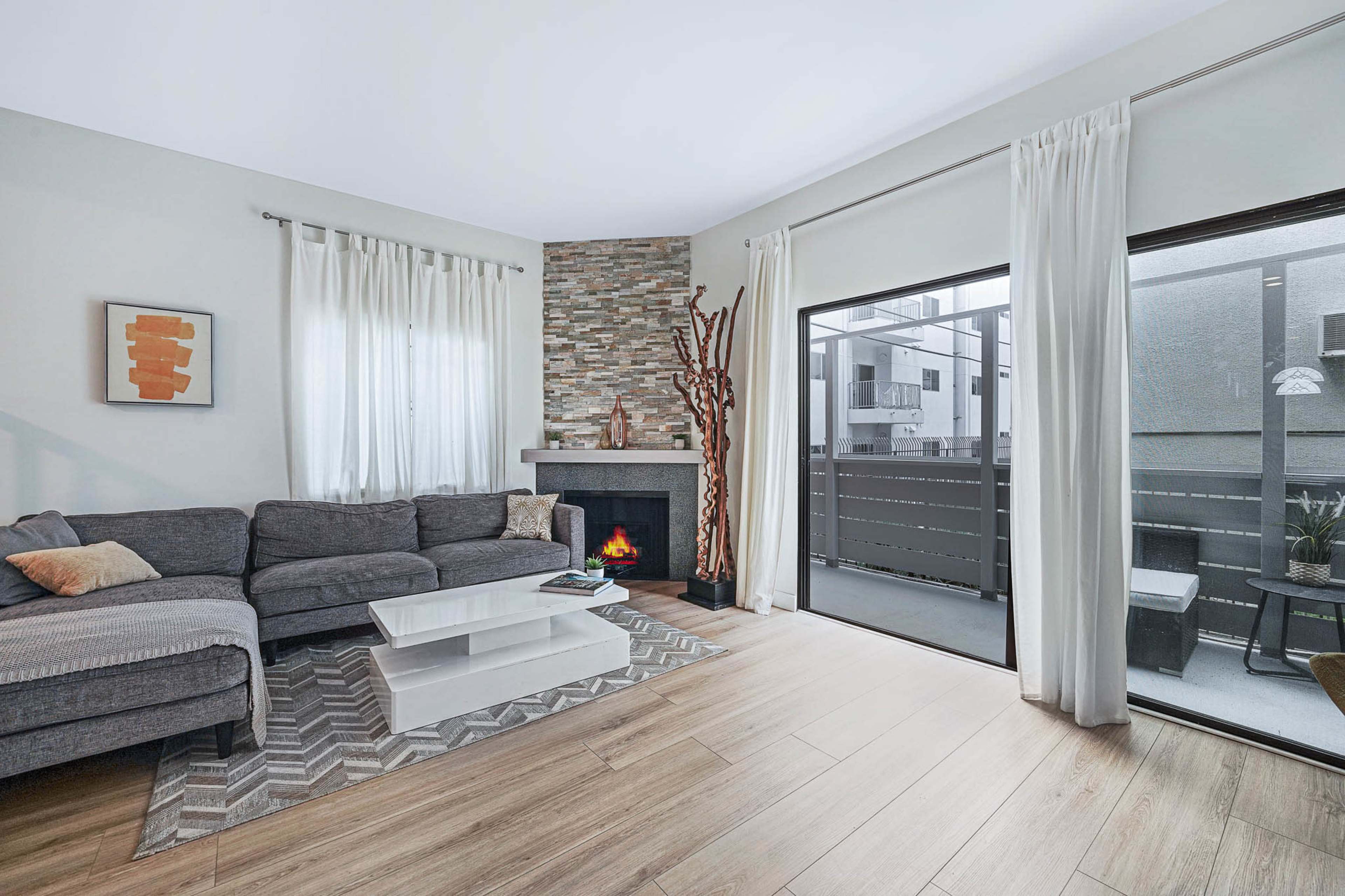 The image features a modern living room with a gray sectional sofa, a stone fireplace, and large windows that open to an outdoor balcony.