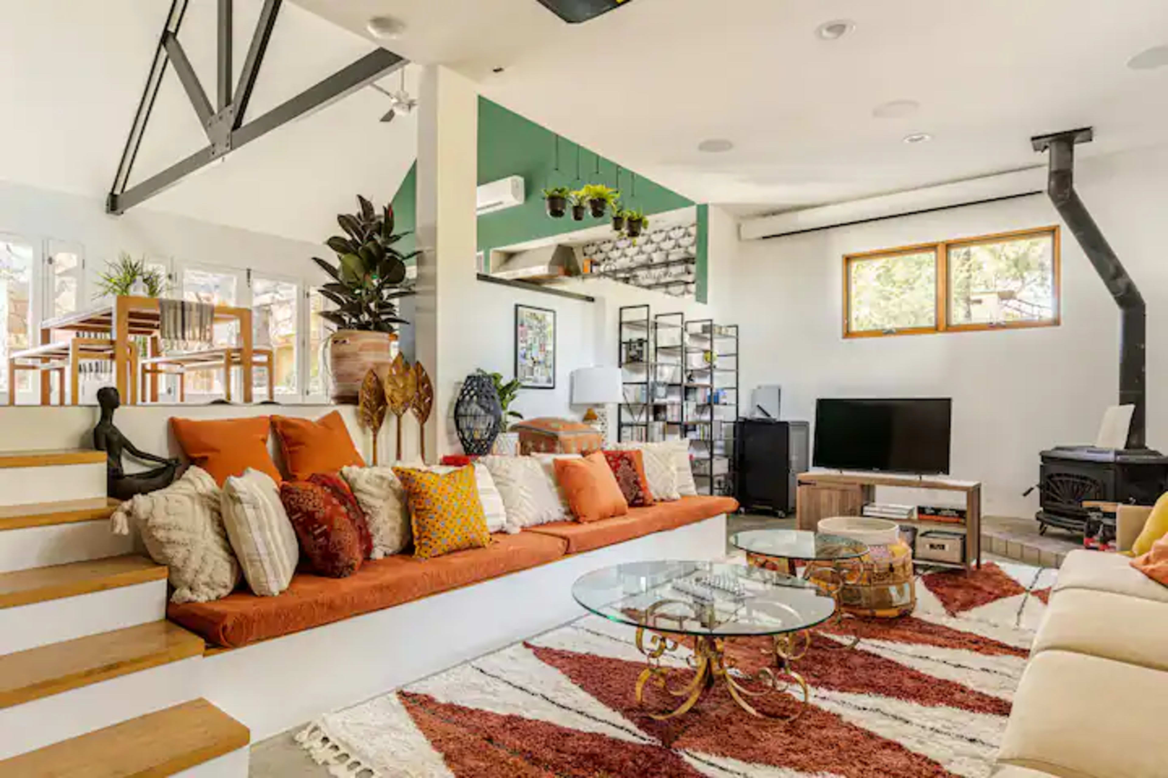 The living room features a sectional sofa with colorful cushions, a glass coffee table, a wood-burning stove, and large windows that provide natural light.
