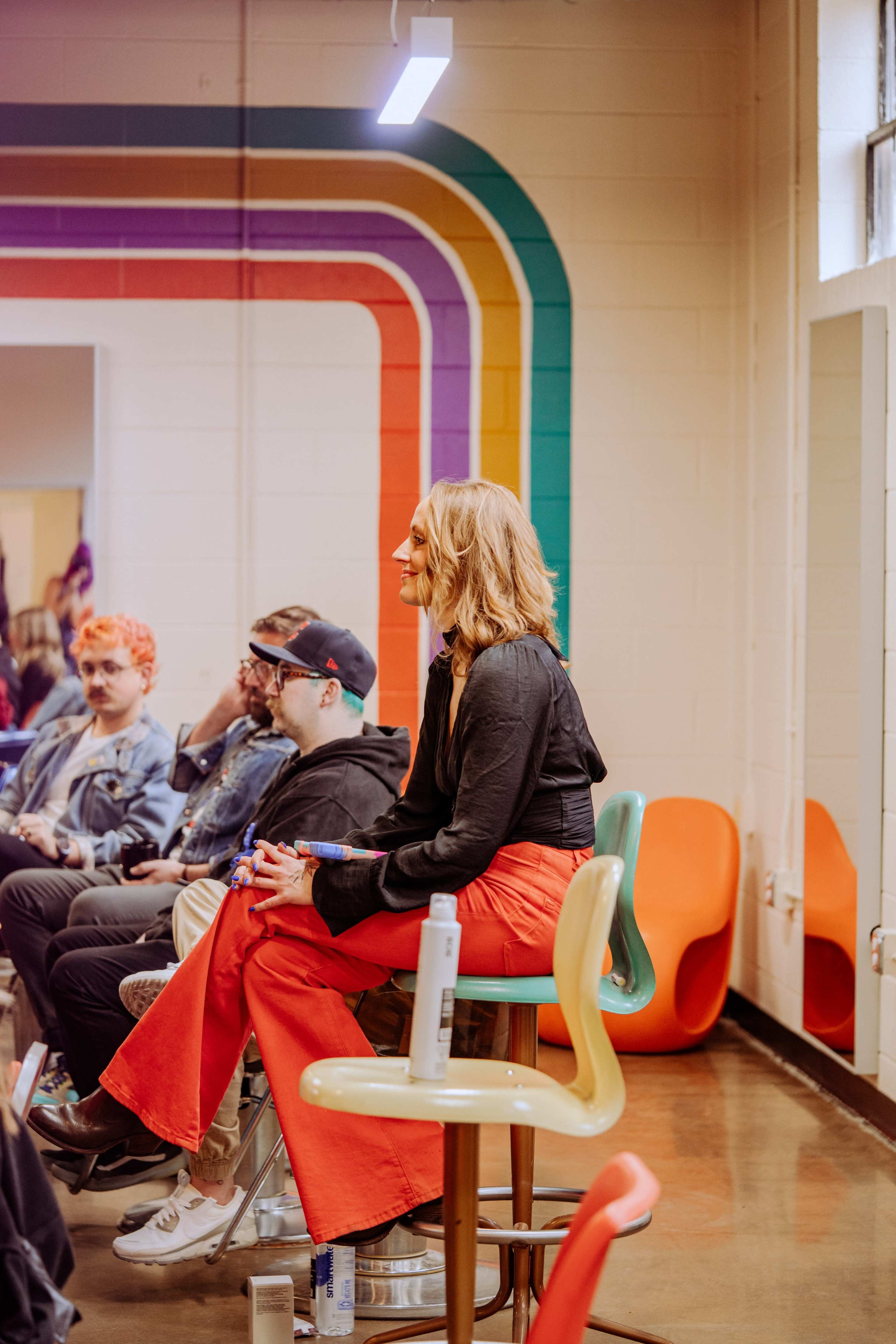 A woman in a black top and red pants sits on a stool, facing a group of people in a colorful, brightly lit room.