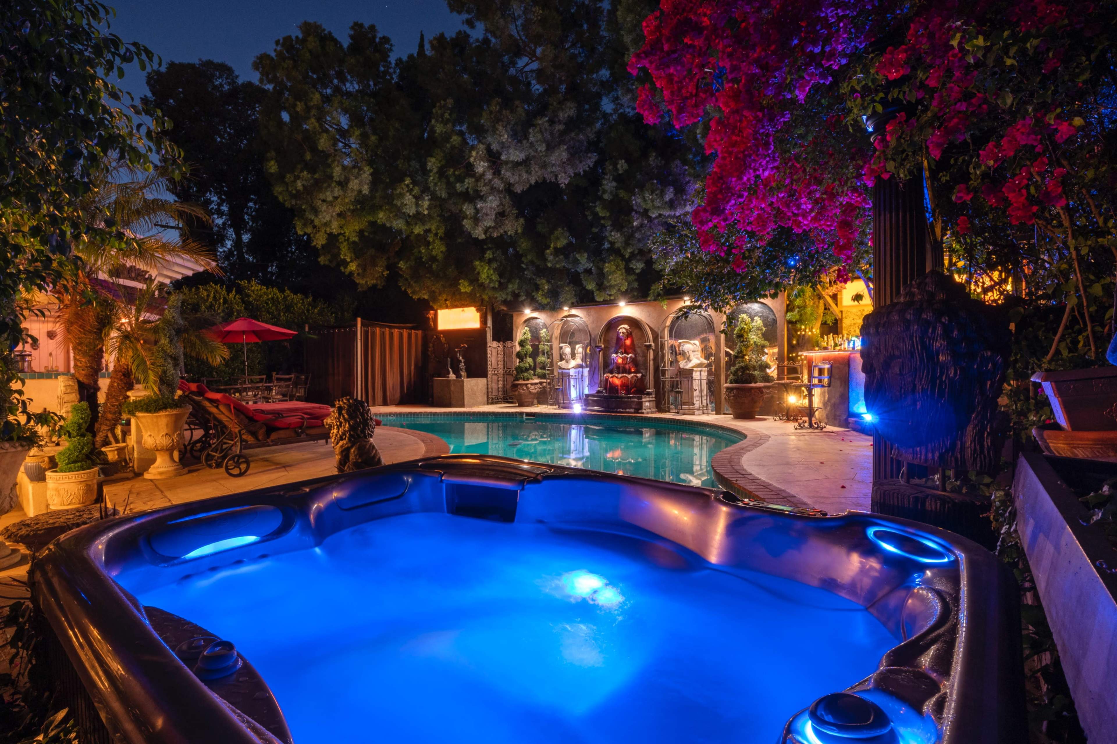 A hot tub with blue lighting is positioned beside a swimming pool surrounded by lush greenery and illuminated architectural features at night.
