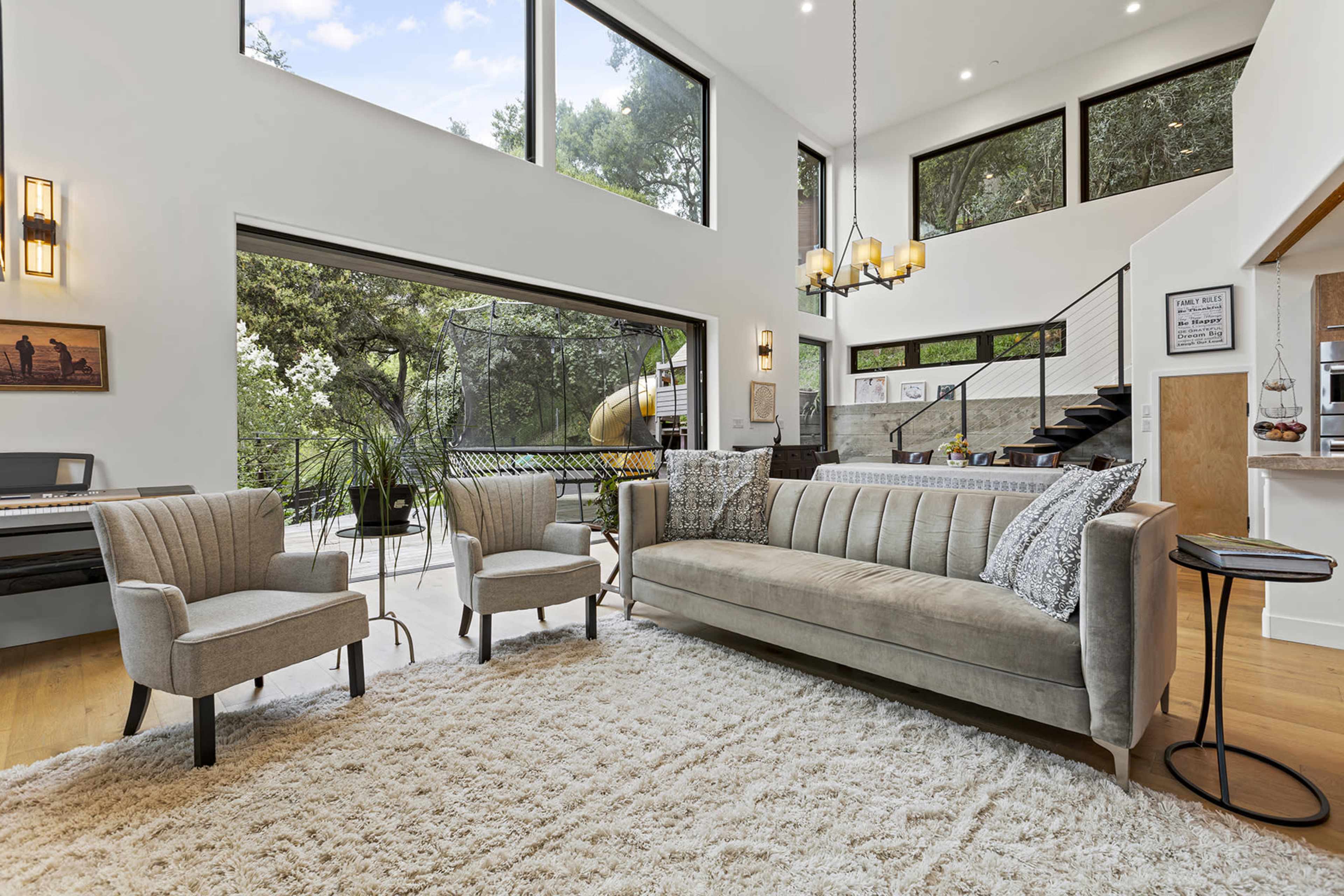 The image shows a modern living room with large windows, a plush sofa, two armchairs, and a staircase leading to an upper level.