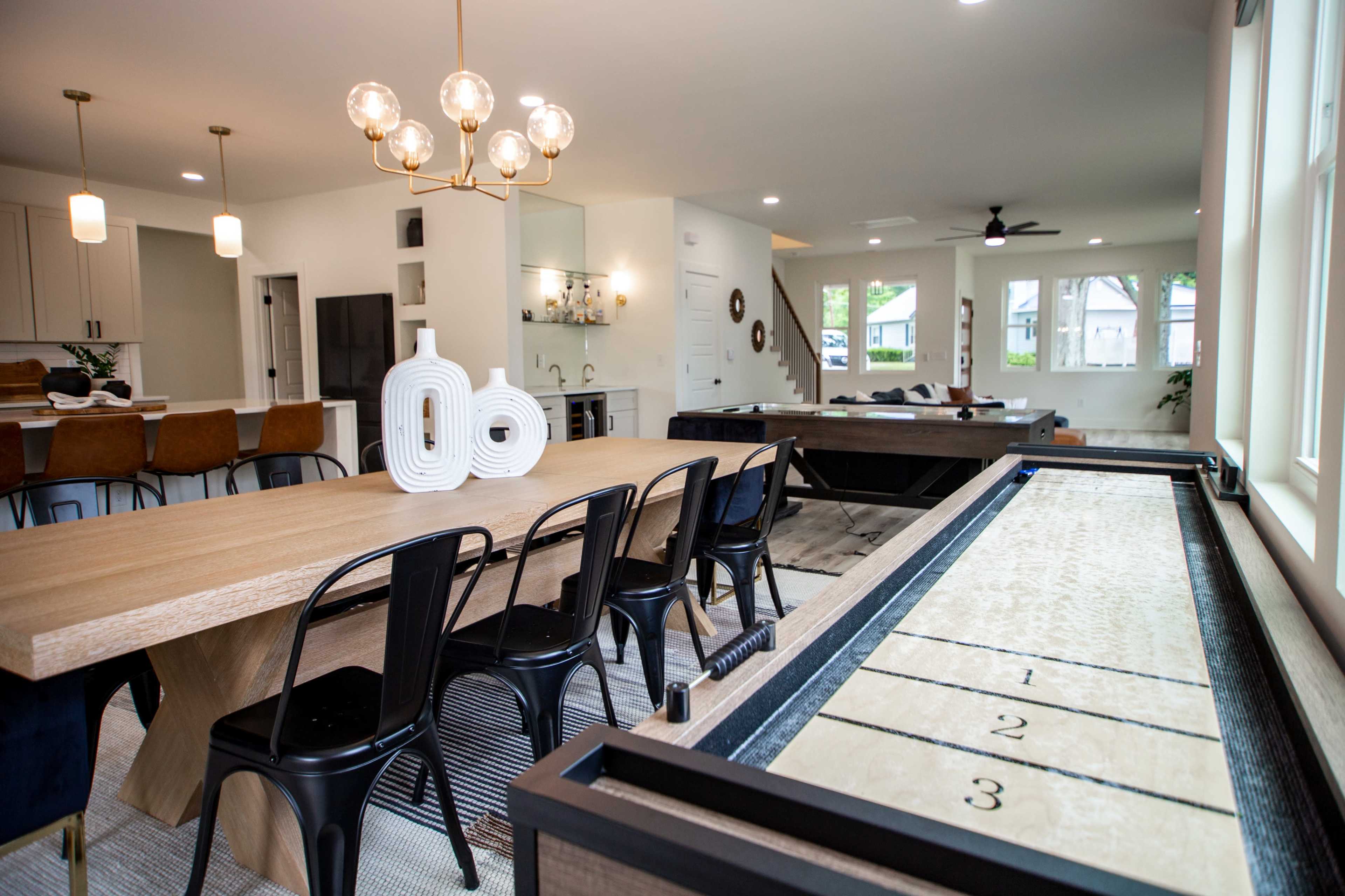 The image shows a modern dining area with a long wooden table surrounded by black chairs, adjacent to a living space featuring a shuffleboard table and large windows.