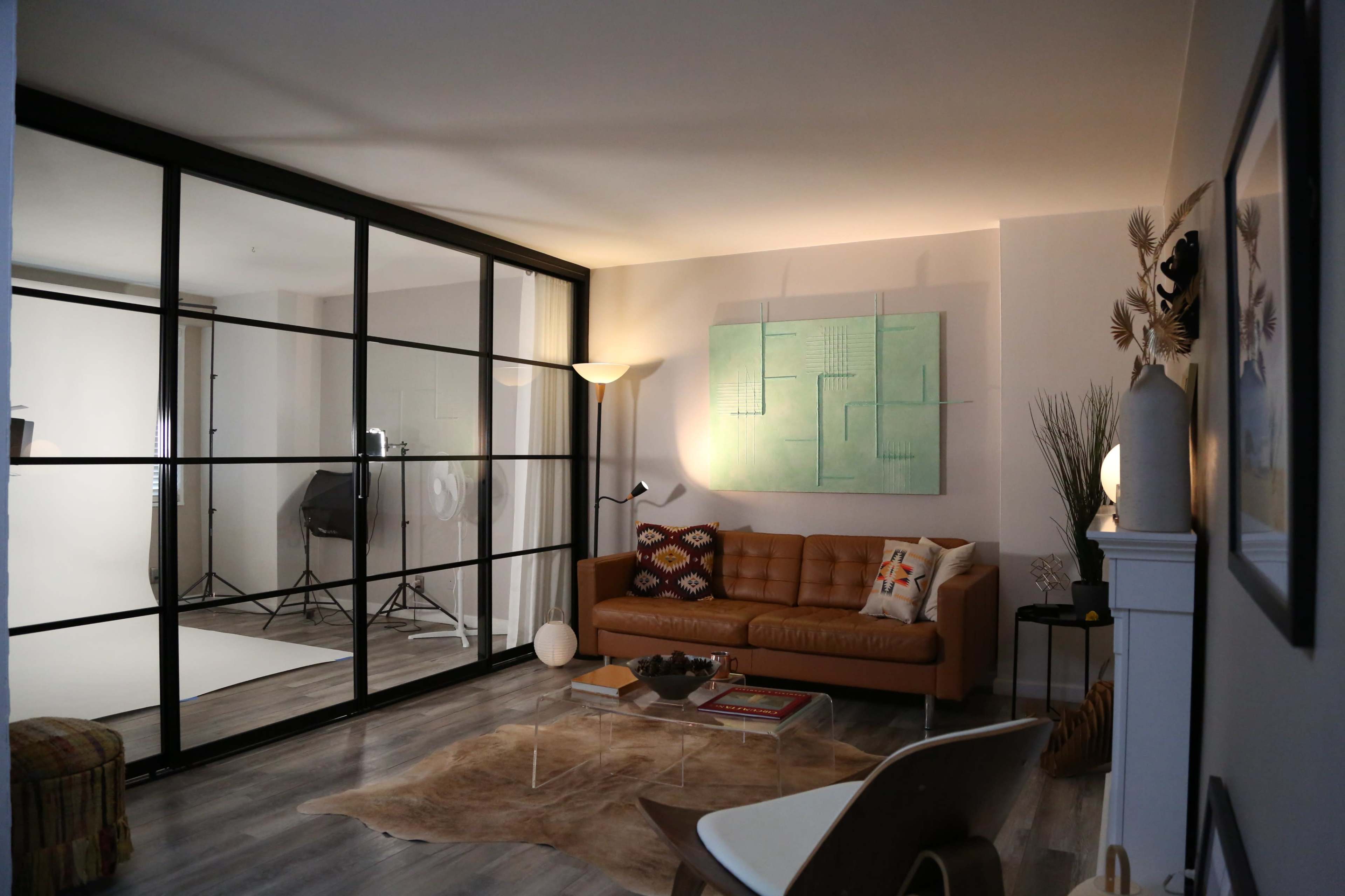 The image shows a contemporary living room with a brown leather sofa, a glass partition, and a soft area rug, illuminated by natural and ambient light.