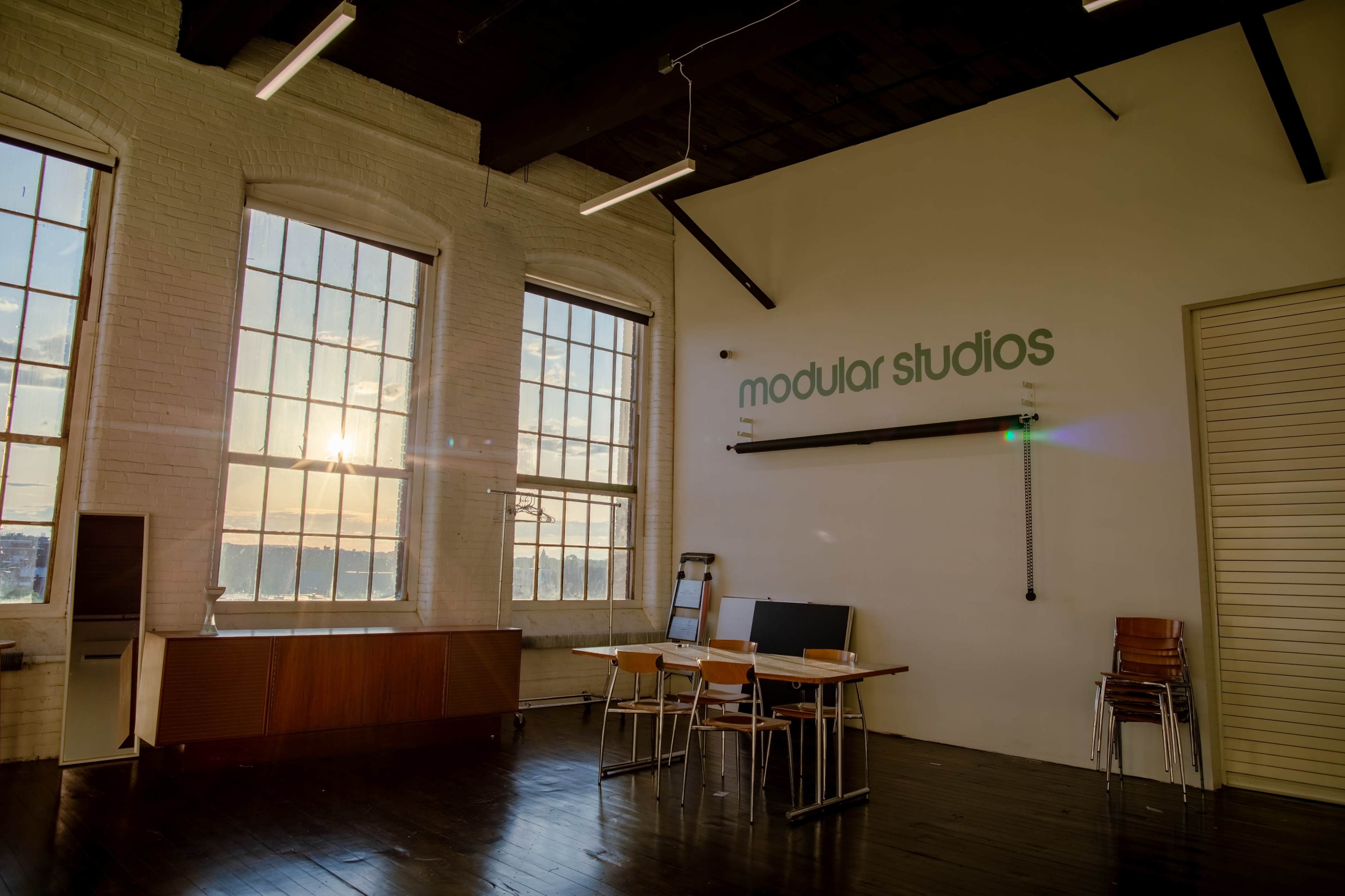A spacious studio with large windows, wooden floors, a table and chairs, and a wall sign that reads "modular studios."