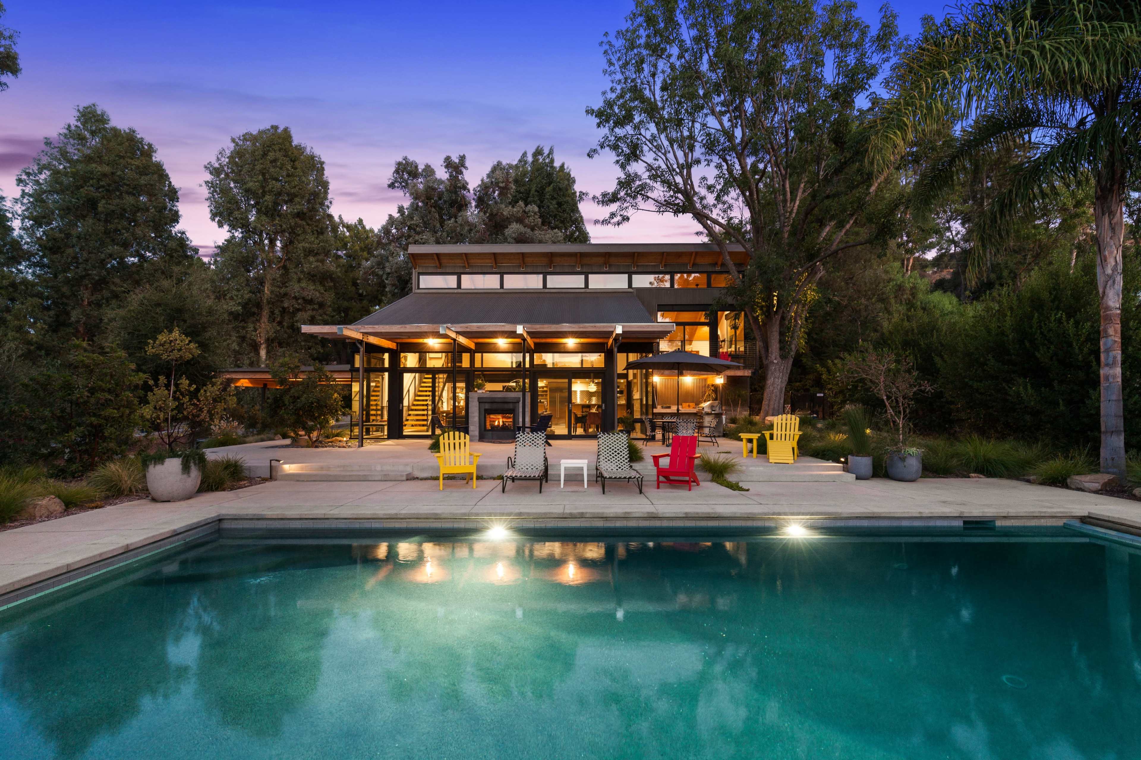 A modern house surrounded by trees features a swimming pool in the foreground and outdoor seating areas with colorful chairs.