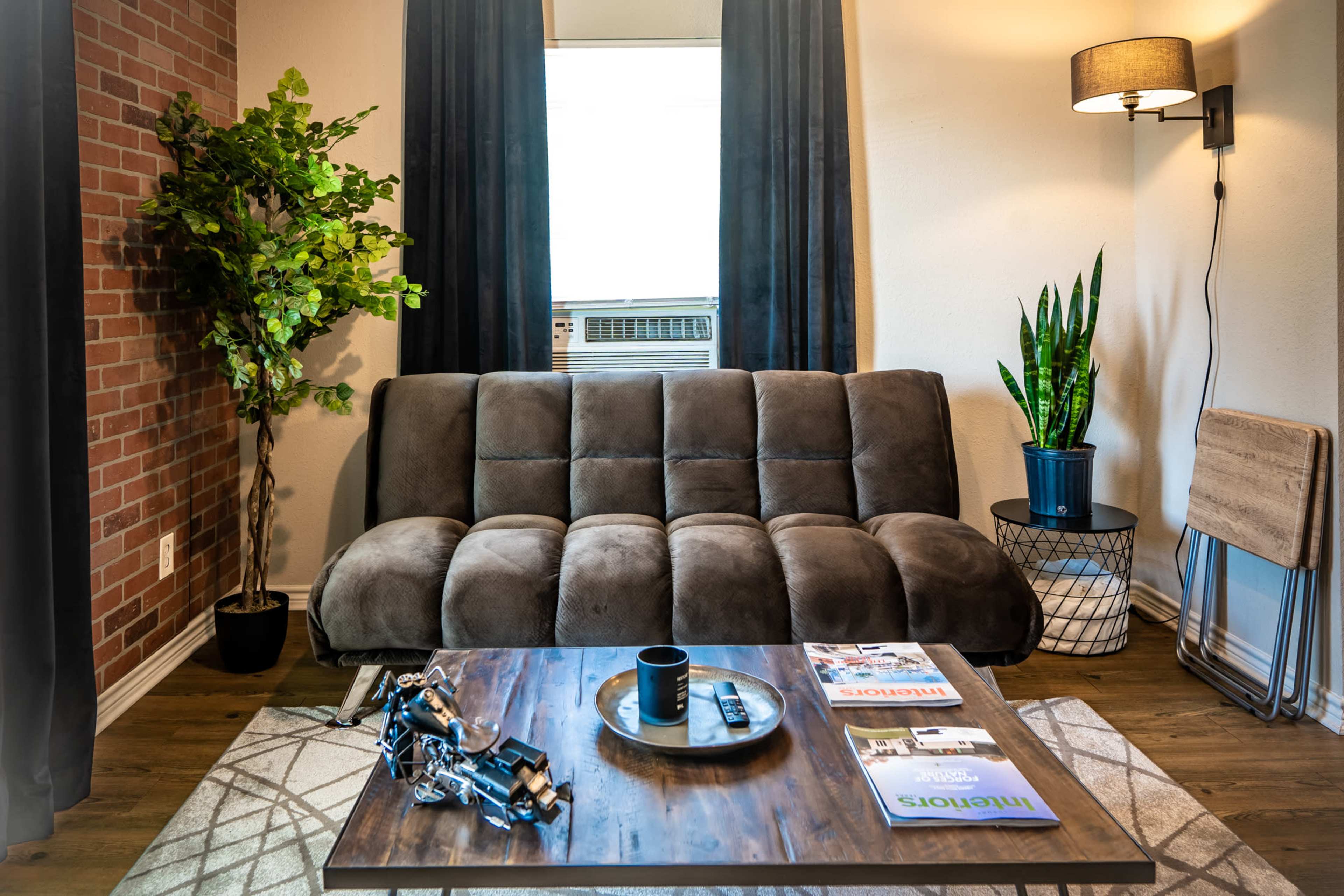 A cozy living room features a gray sofa, a coffee table with magazines and a remote, potted plants, and an air conditioning unit by the window.