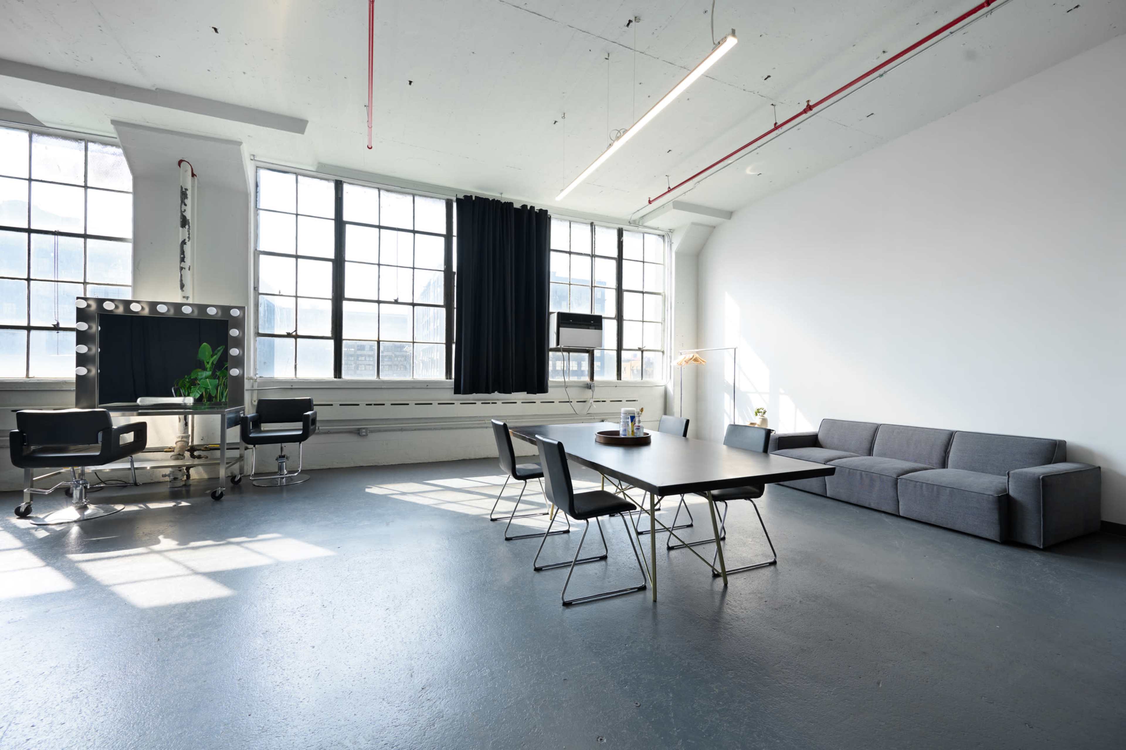 A spacious, modern office interior with large windows, a table surrounded by chairs, a sofa, and a makeup station with mirrors.