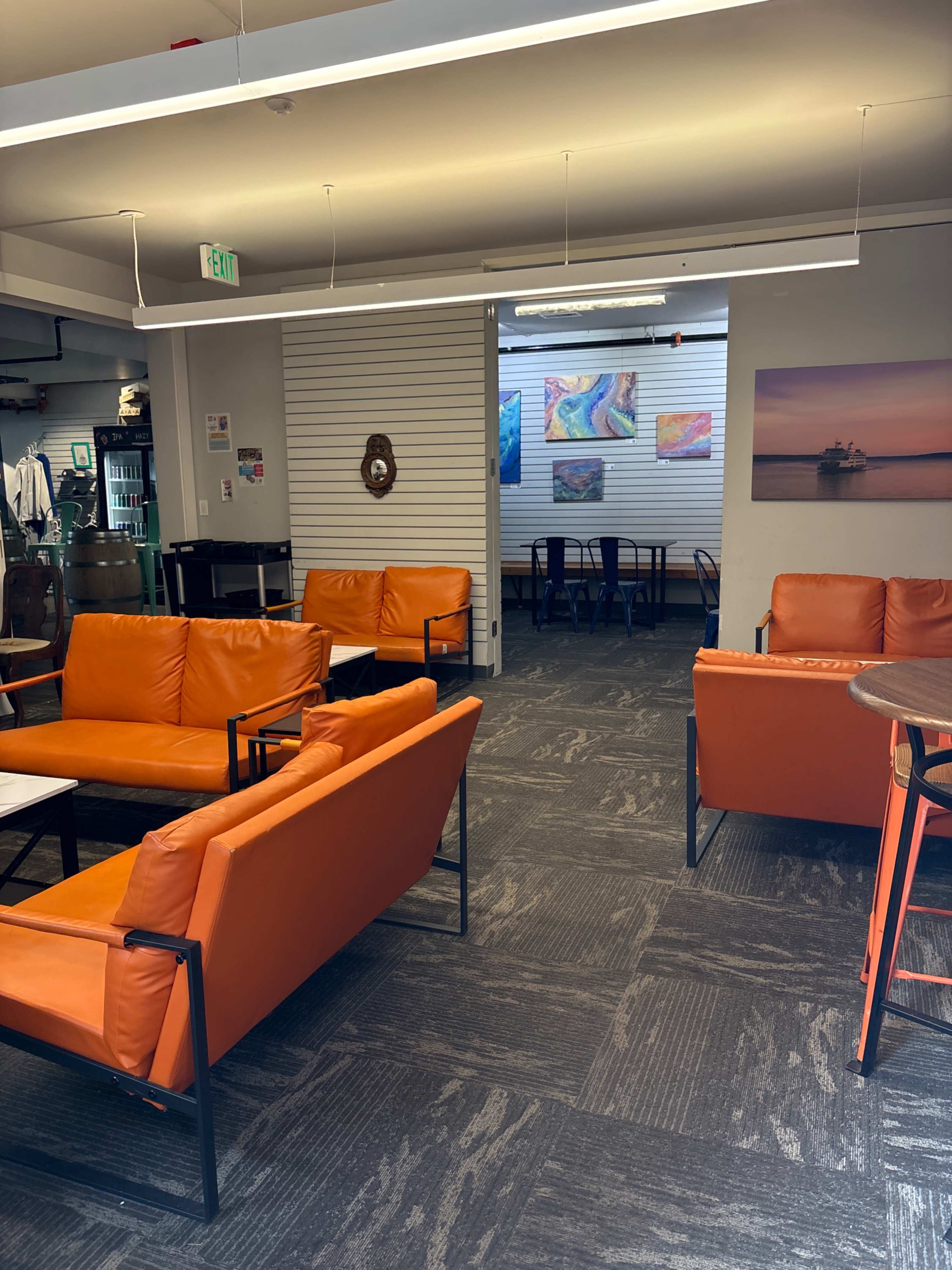 The image shows a spacious room featuring several orange seating arrangements, a few tables, and colorful artwork on the walls.