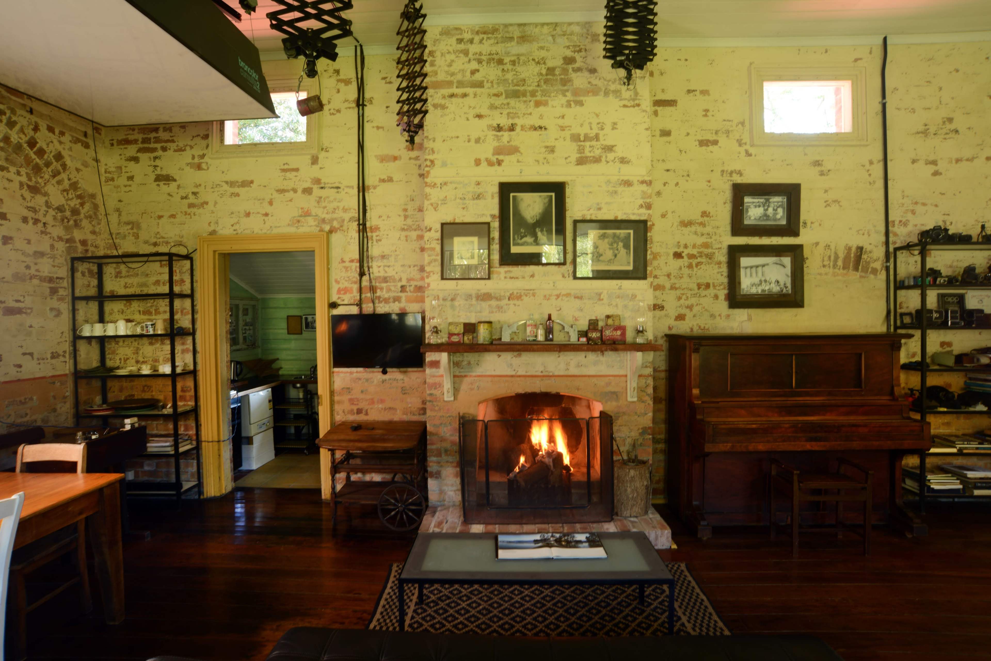 A rustic living room with a brick wall, a fireplace with a fire burning, a piano, and various framed pictures on the wall.