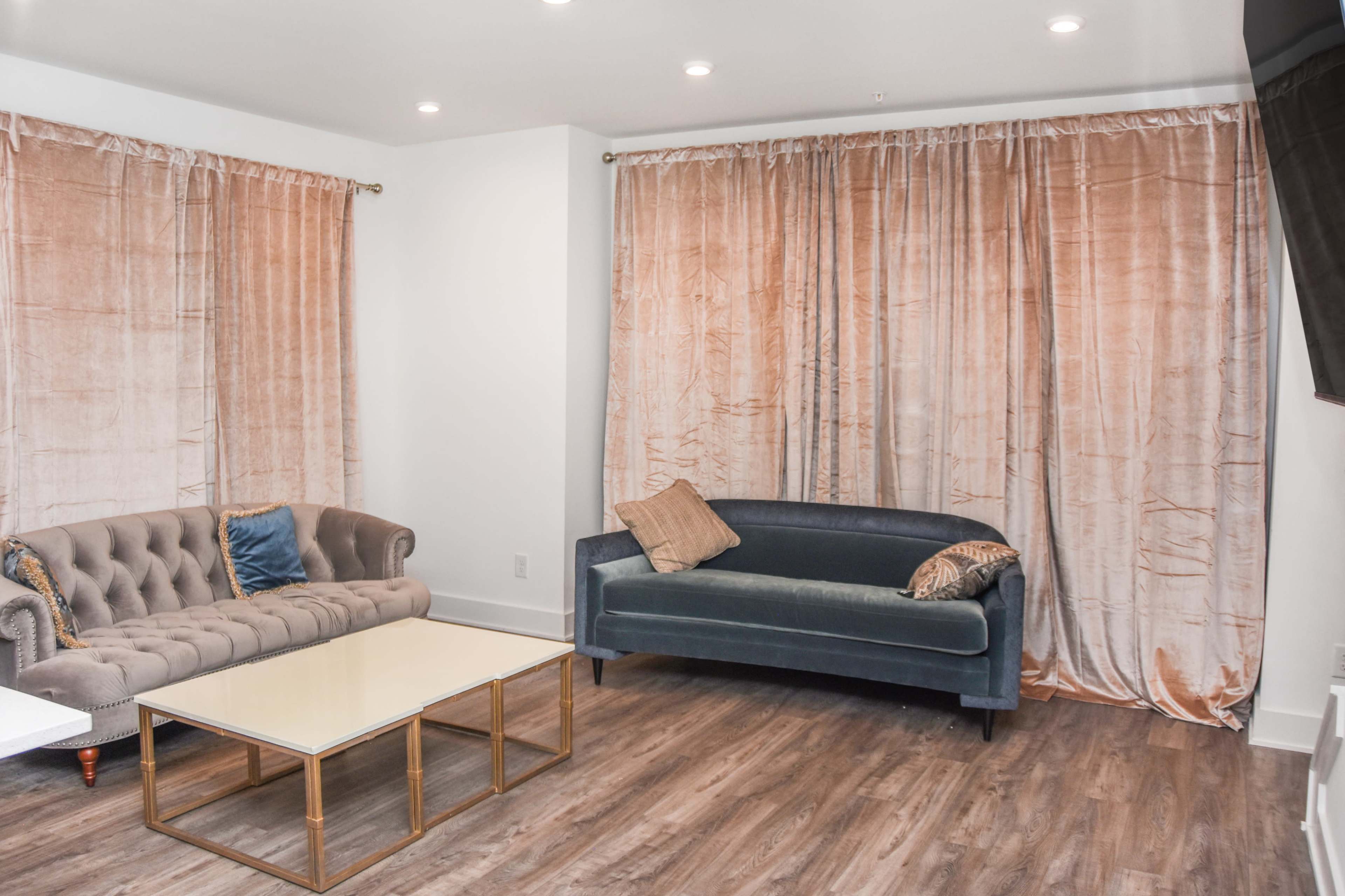The image shows a living room with a light-colored wall, two elegant sofas, a coffee table, and floor-to-ceiling curtains.