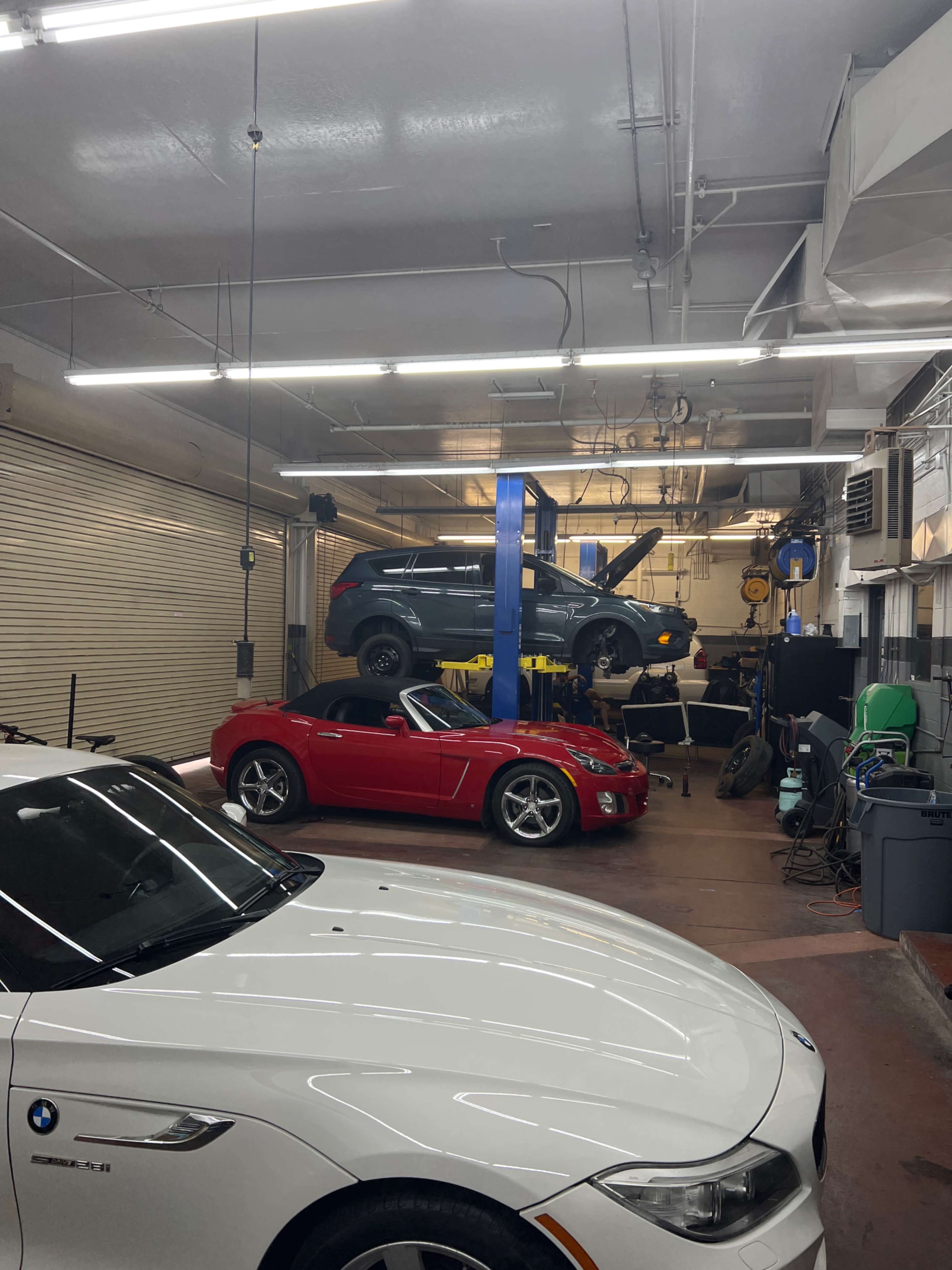 The image shows a car repair shop with a red convertible in the foreground, a white car on the left, and a blue vehicle being serviced on a lift in the background.