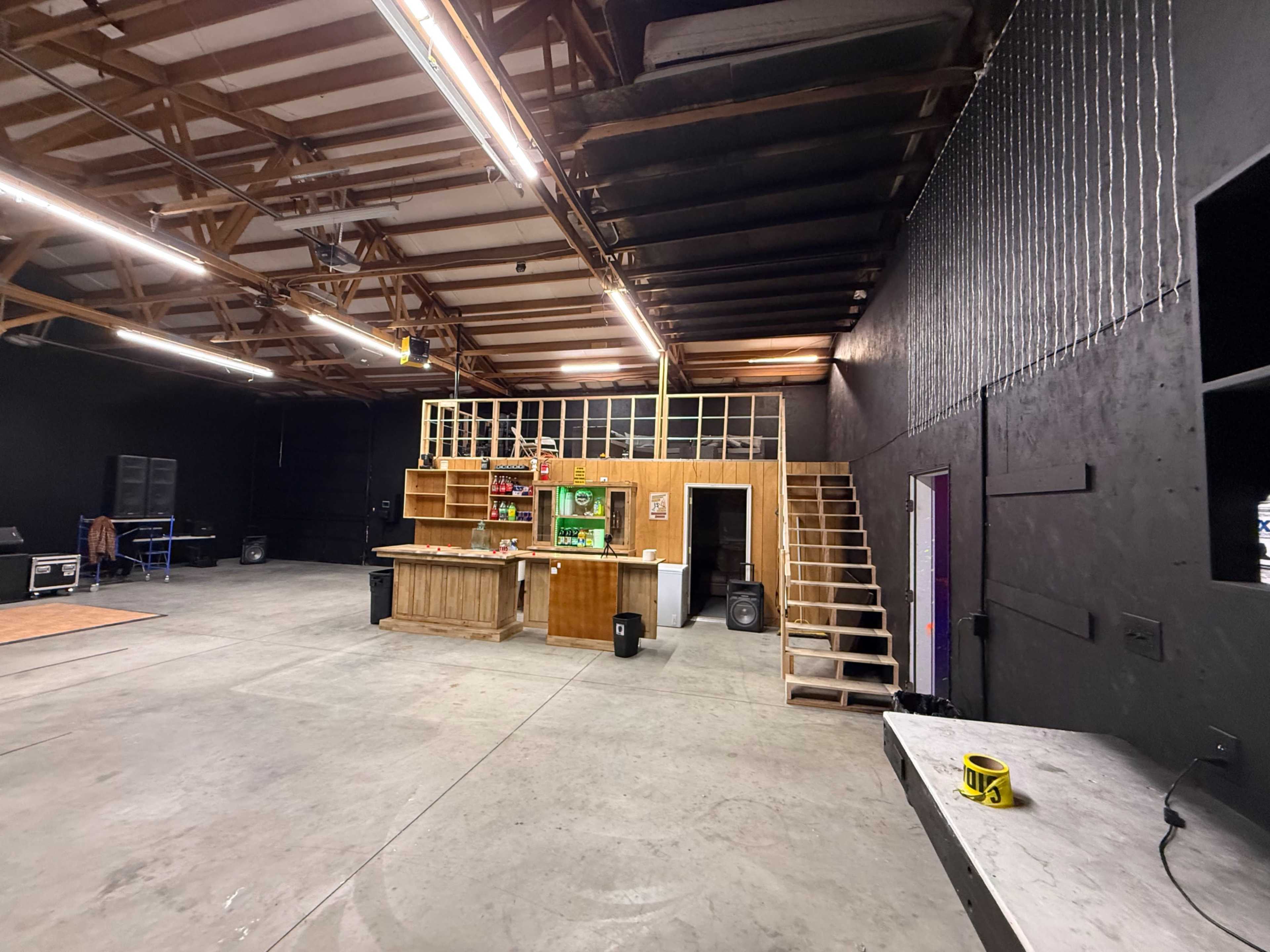 The image shows a large, empty studio space with a wooden bar and a staircase leading to an upper level against a black wall.
