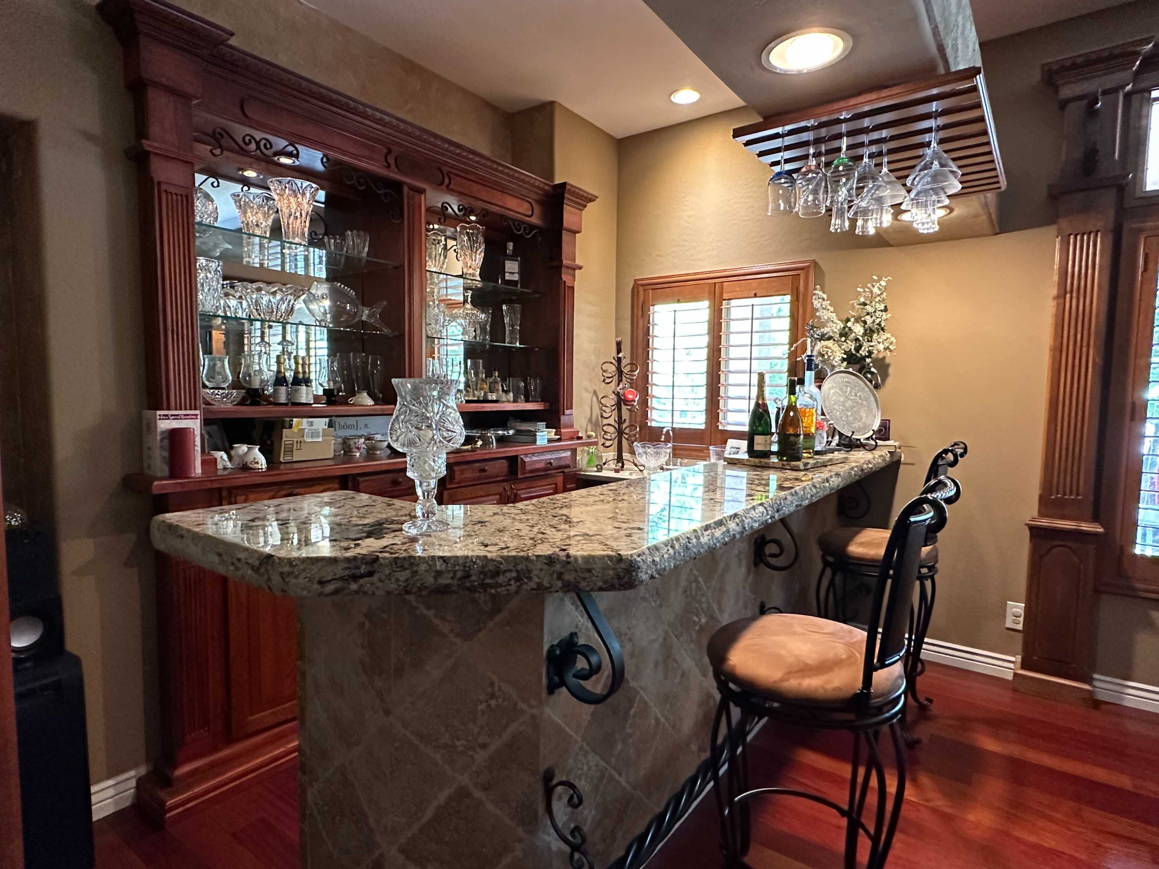 A home bar area with a granite countertop, wooden cabinetry, and glassware displayed on shelves, complemented by bar stools and a decorative arrangement.