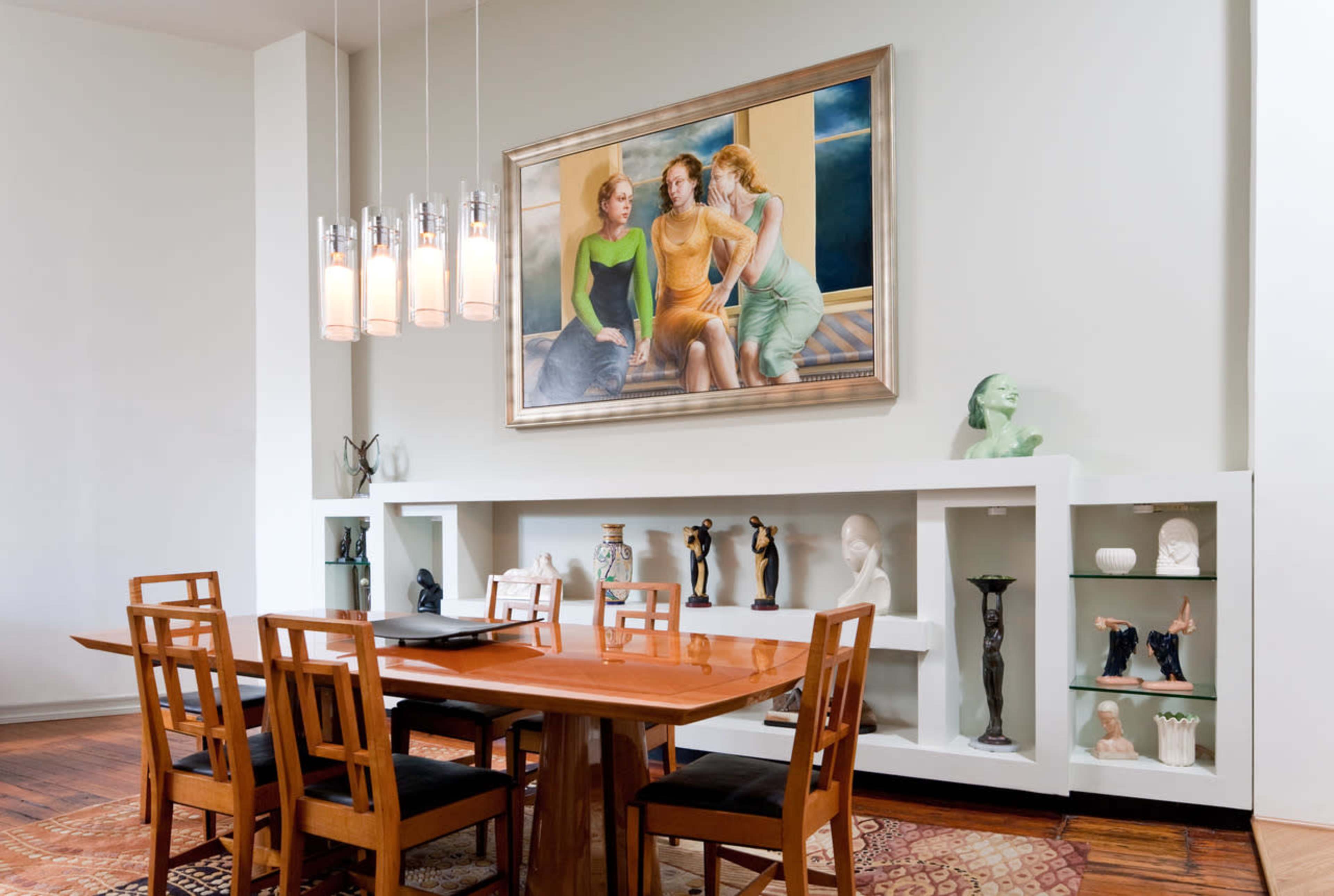 A dining area features a wooden table surrounded by chairs, with a prominent painting of three women on the wall and a display of sculptures on shelves.