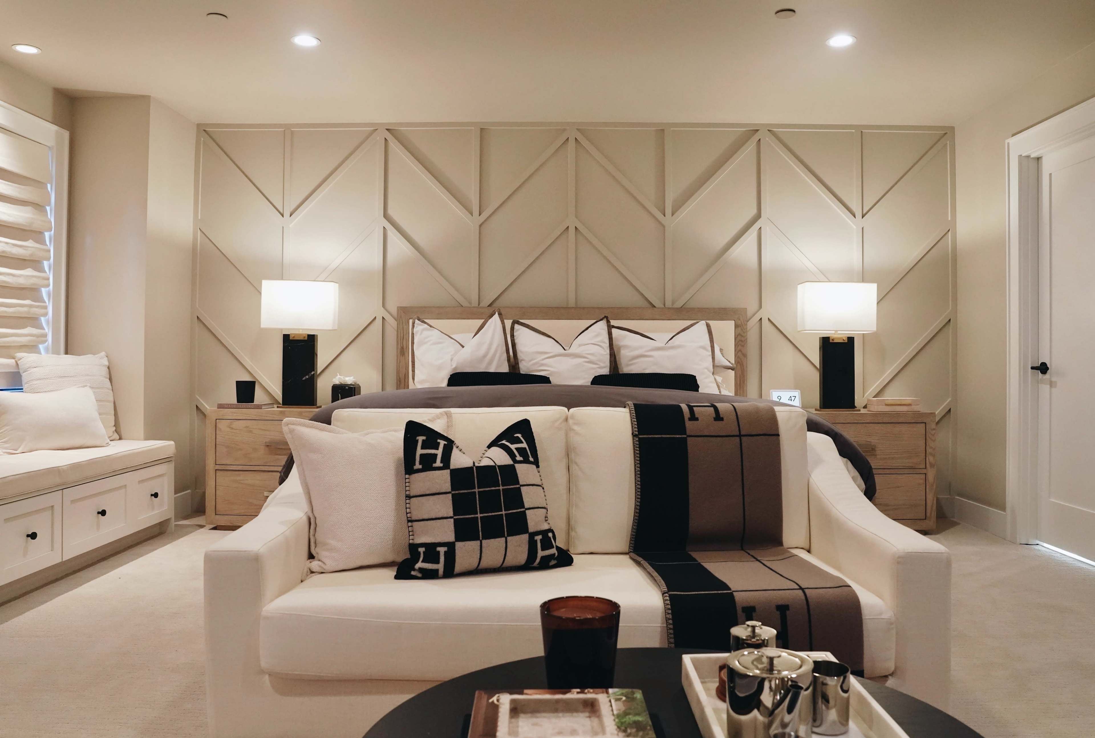 A cozy bedroom features a modern design with a light-colored couch adorned with decorative pillows, a large bed in the background, and bedside lamps.