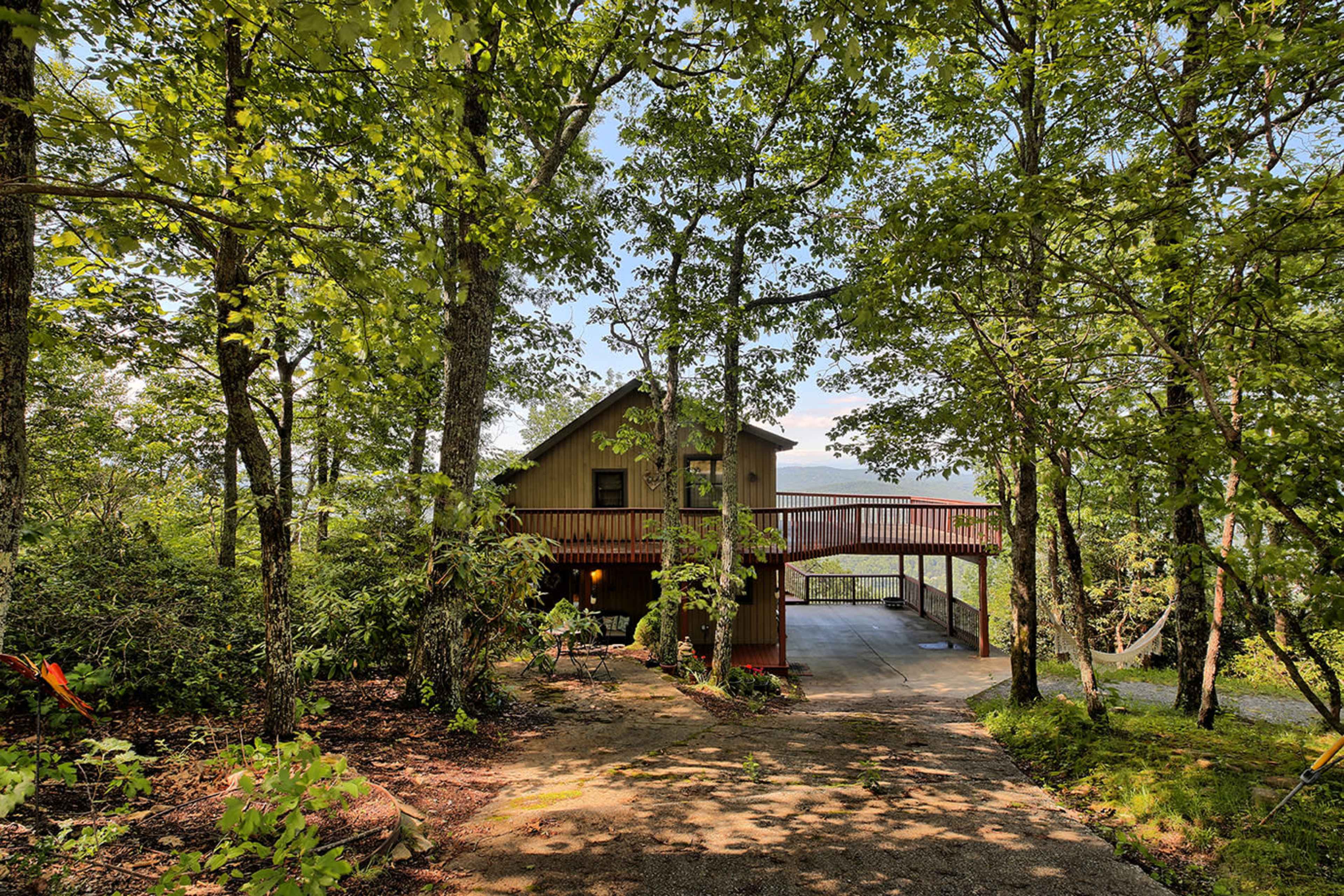 A two-story house with a wooden deck is located among trees on a sloped driveway.