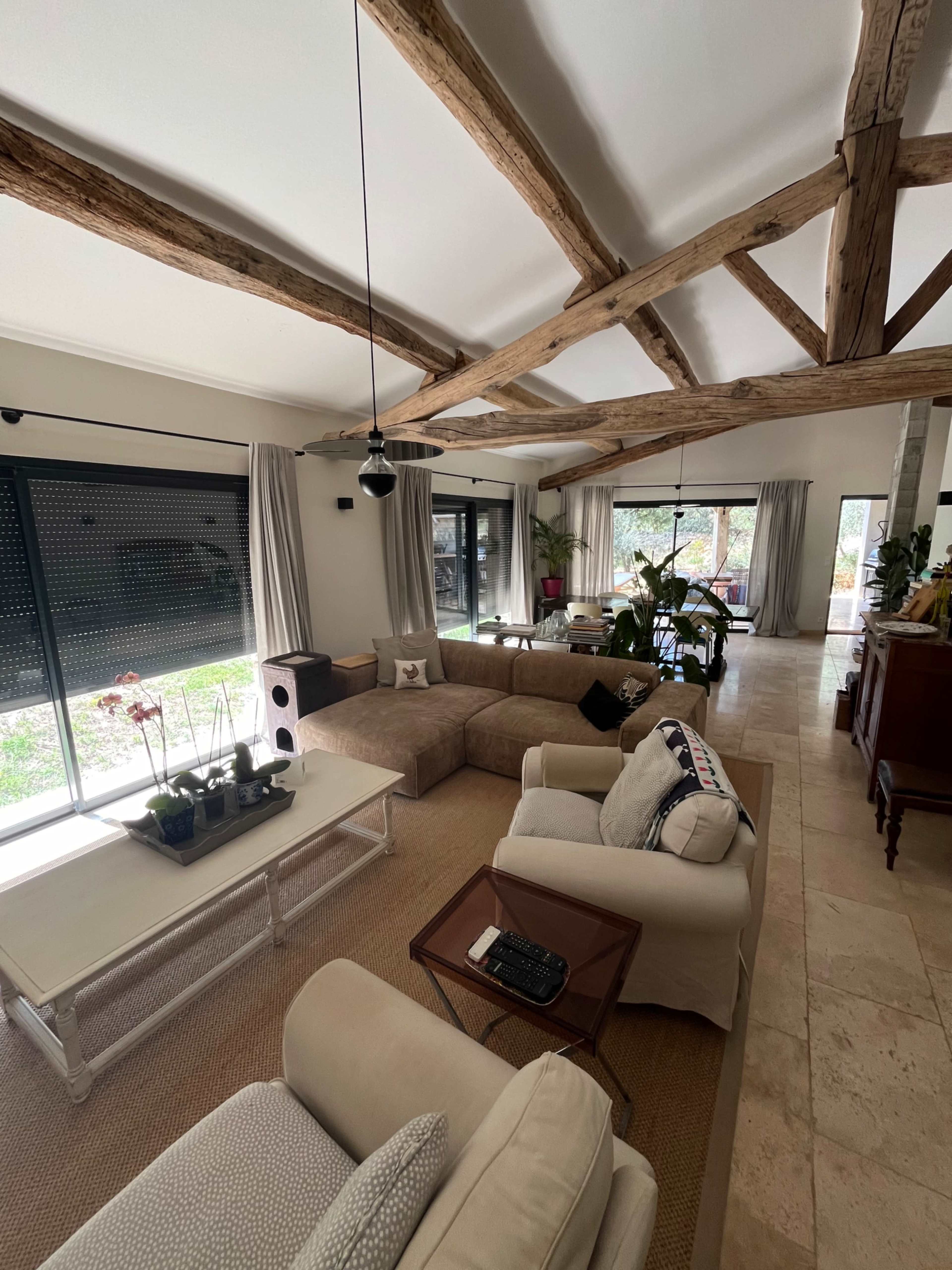 The living room features a large sectional sofa, a wooden coffee table, and exposed wooden beams on the ceiling, with large windows overlooking a garden.