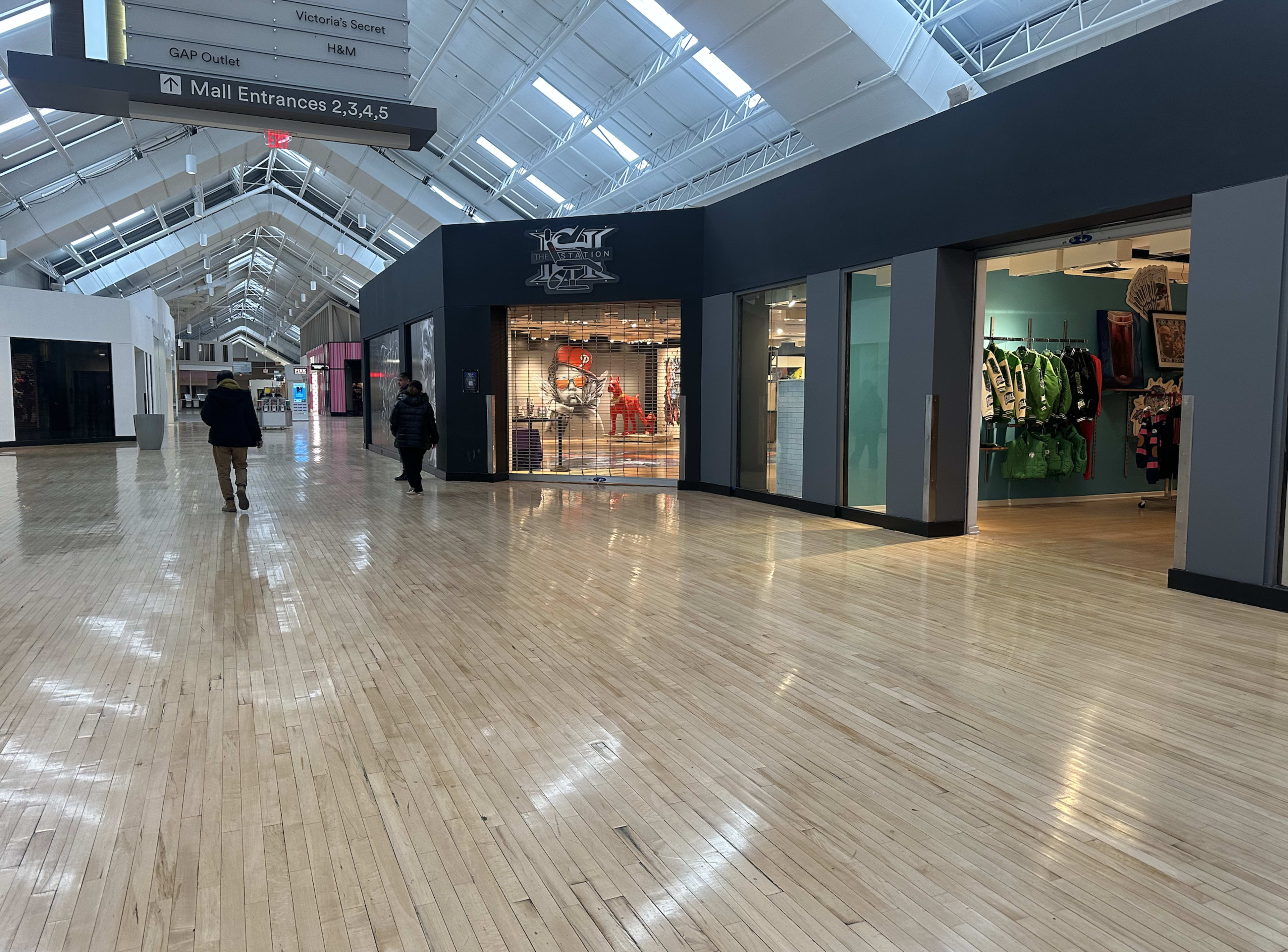 The image shows an indoor mall corridor with storefronts on either side and people walking through the space.