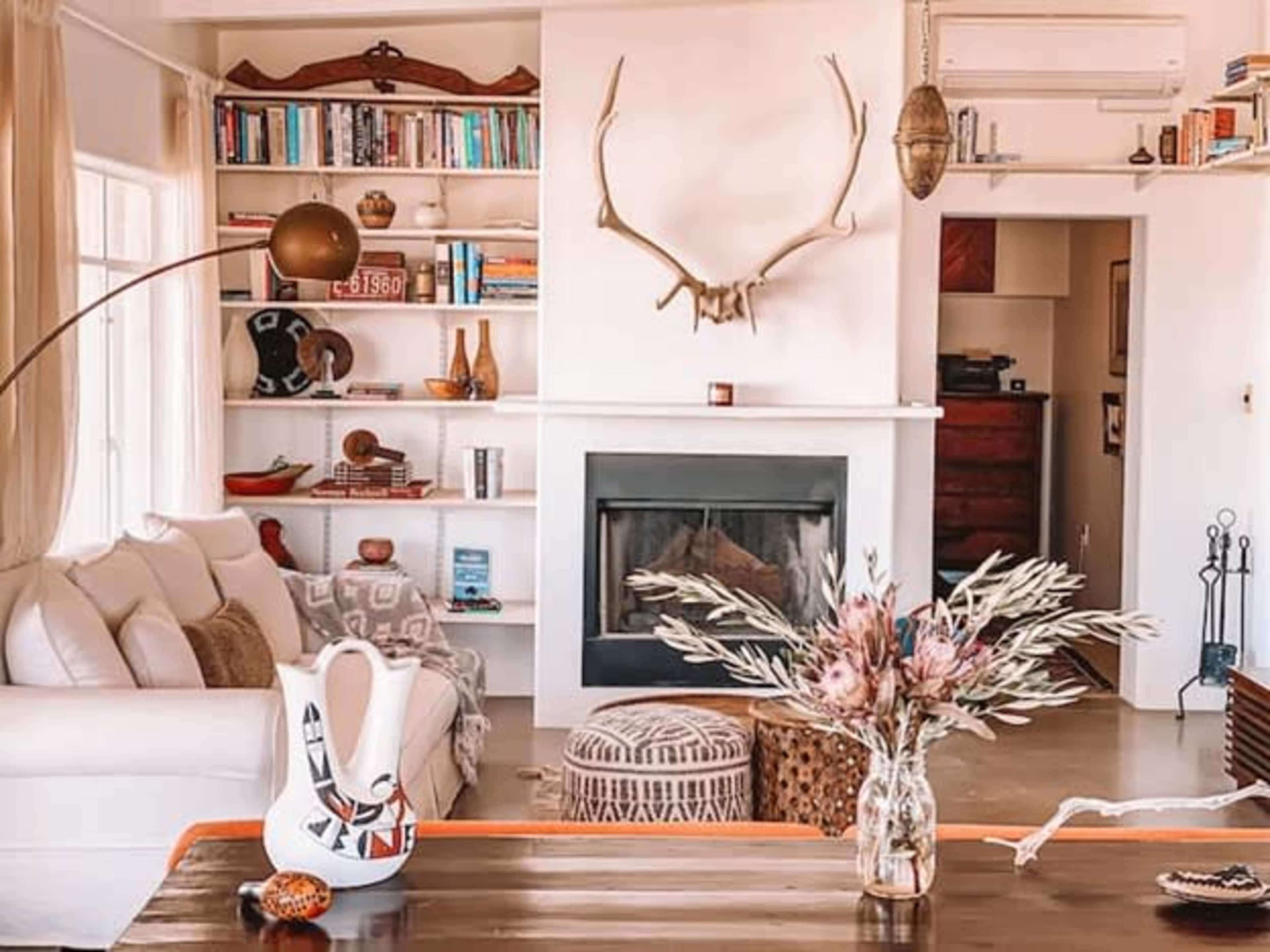 A spacious living room features a white sofa, a fireplace with a simple mantle, bookshelves filled with books, and decorative elements like an antler and a vase of dried flowers.