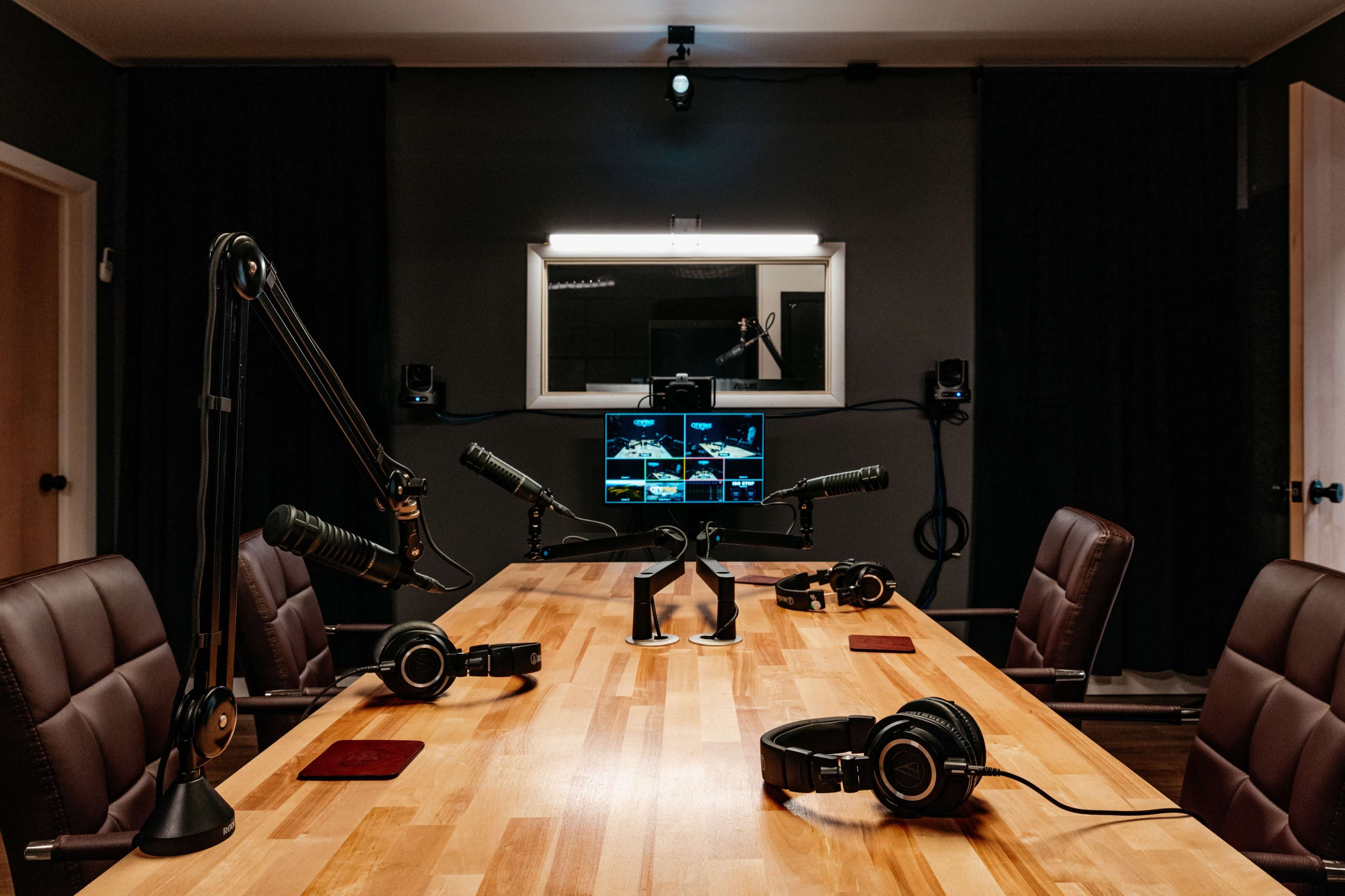 A modern podcasting studio features a large wooden table with multiple microphones, headphones, and a computer monitor displaying various audio tracks.