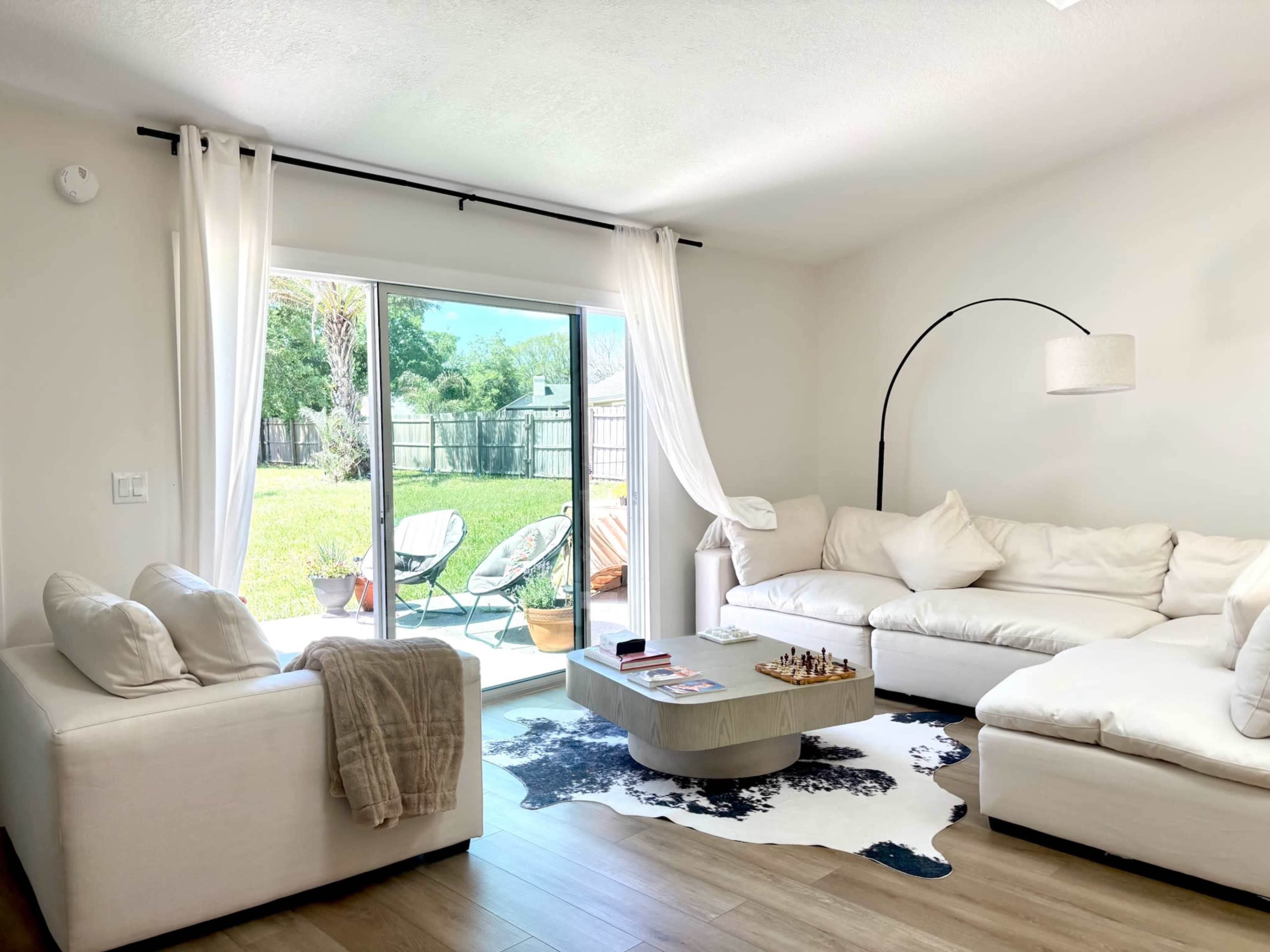 The image shows a bright living room featuring a white sectional sofa, a matching armchair, a coffee table on a cowhide rug, and large sliding glass doors leading to a grassy outdoor area.