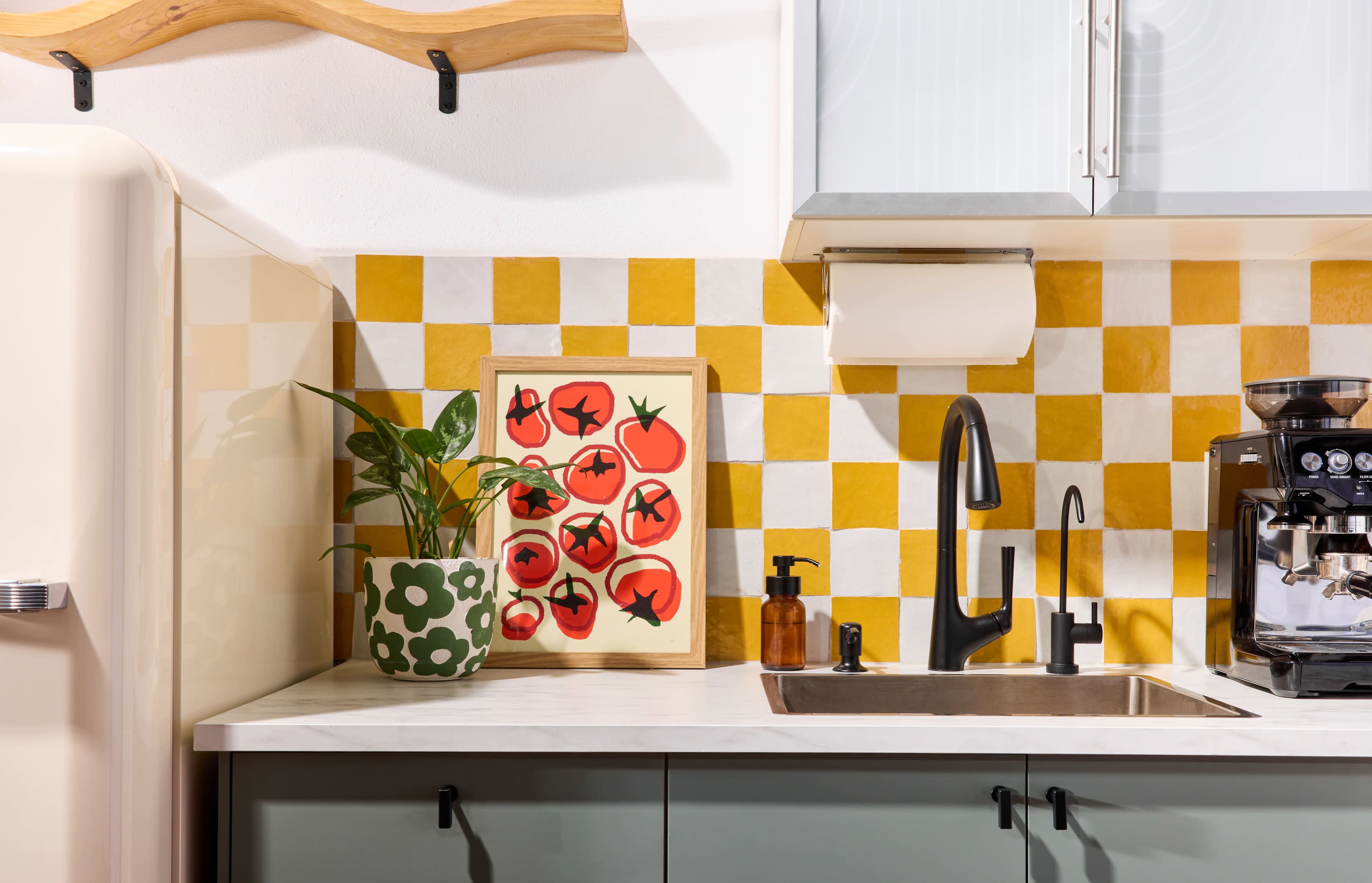 The image shows a modern kitchen with yellow and white checkered tiles, featuring a plant, a framed artwork of tomatoes, a sink, and a coffee machine.