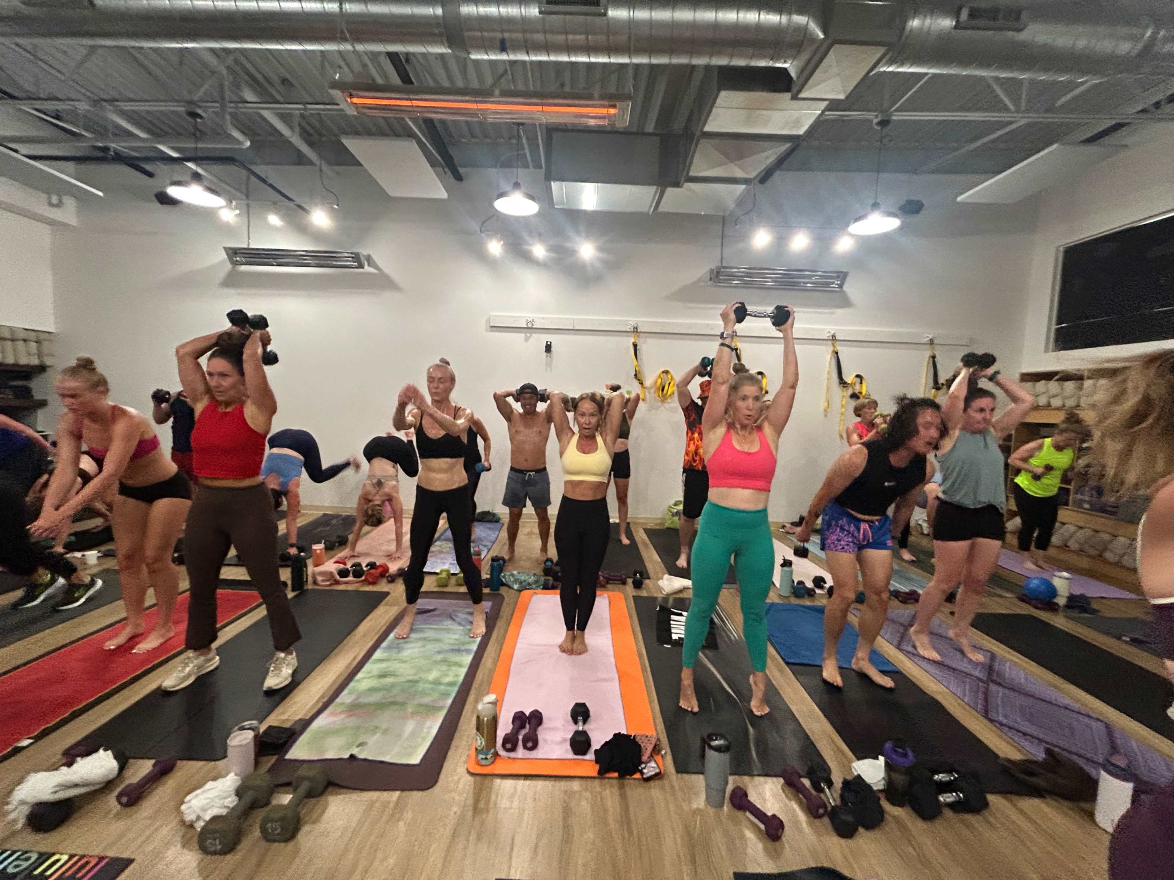 A group of people in workout attire are engaged in an exercise class, using dumbbells and yoga mats in a well-lit gym.
