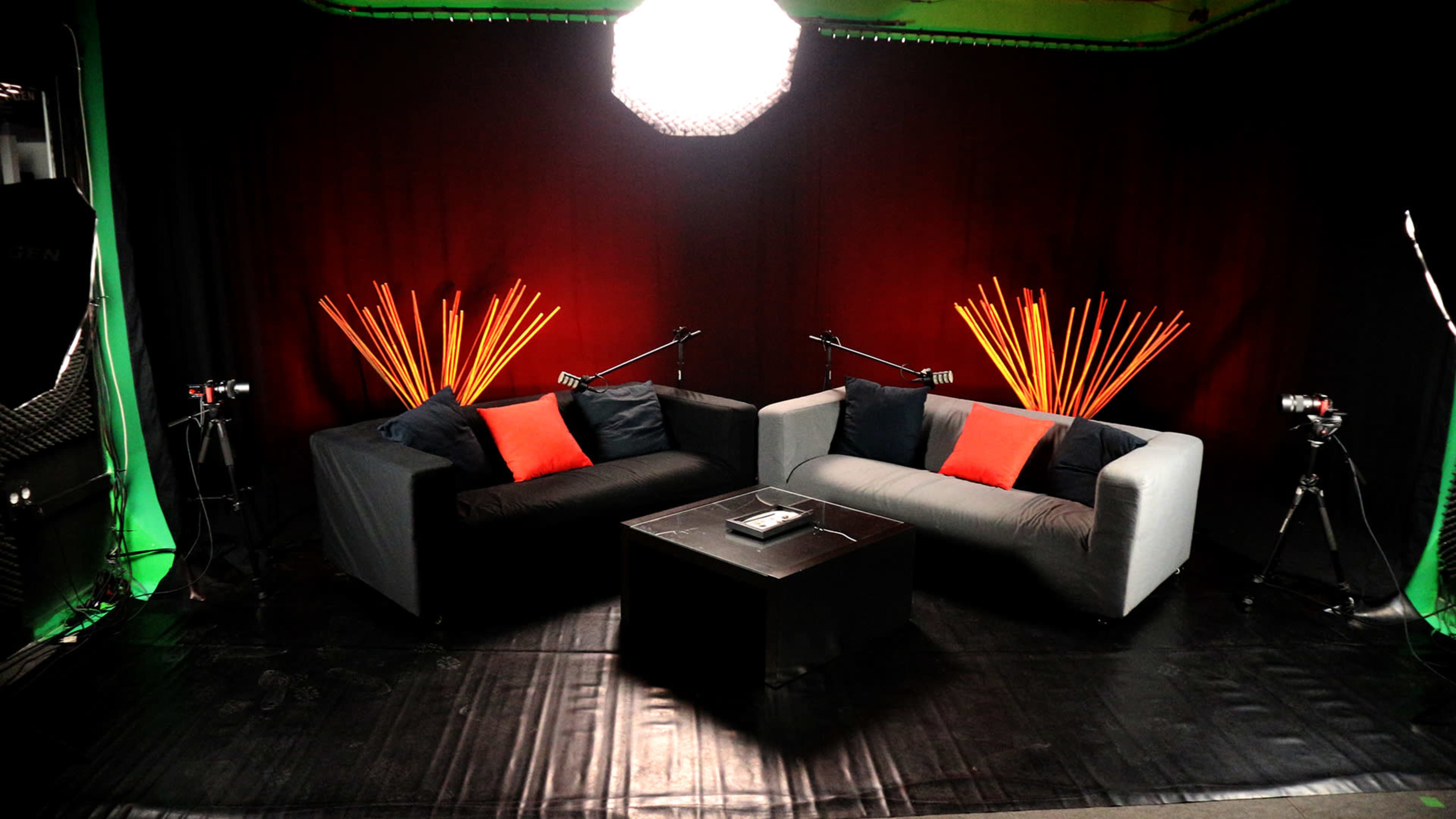 The image shows a television studio set with two sofas, a coffee table, and lighting equipment, framed by a dark red backdrop.