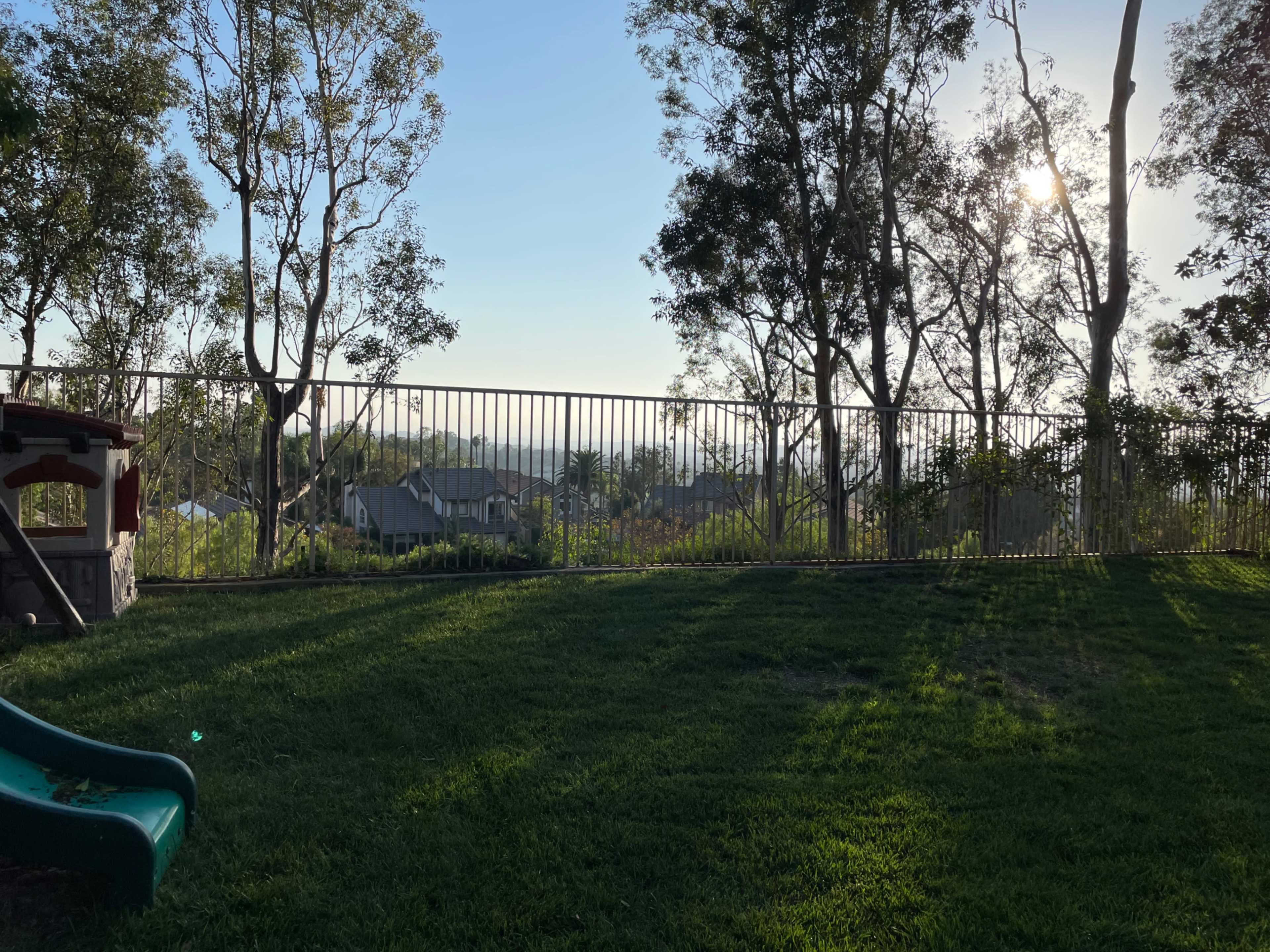A fenced backyard with green grass and a play structure, surrounded by trees and a distant view of houses and hills.