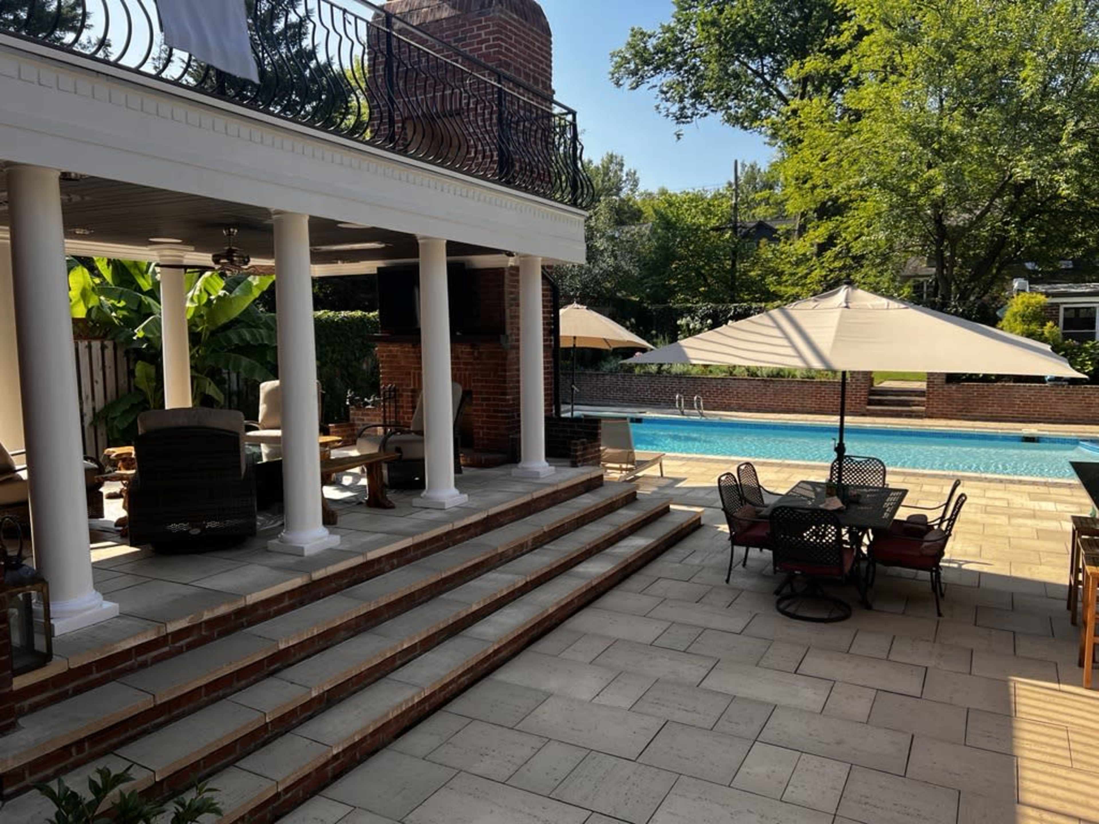 A patio area with steps leading to a swimming pool, surrounded by lounge chairs and dining tables beneath umbrellas.