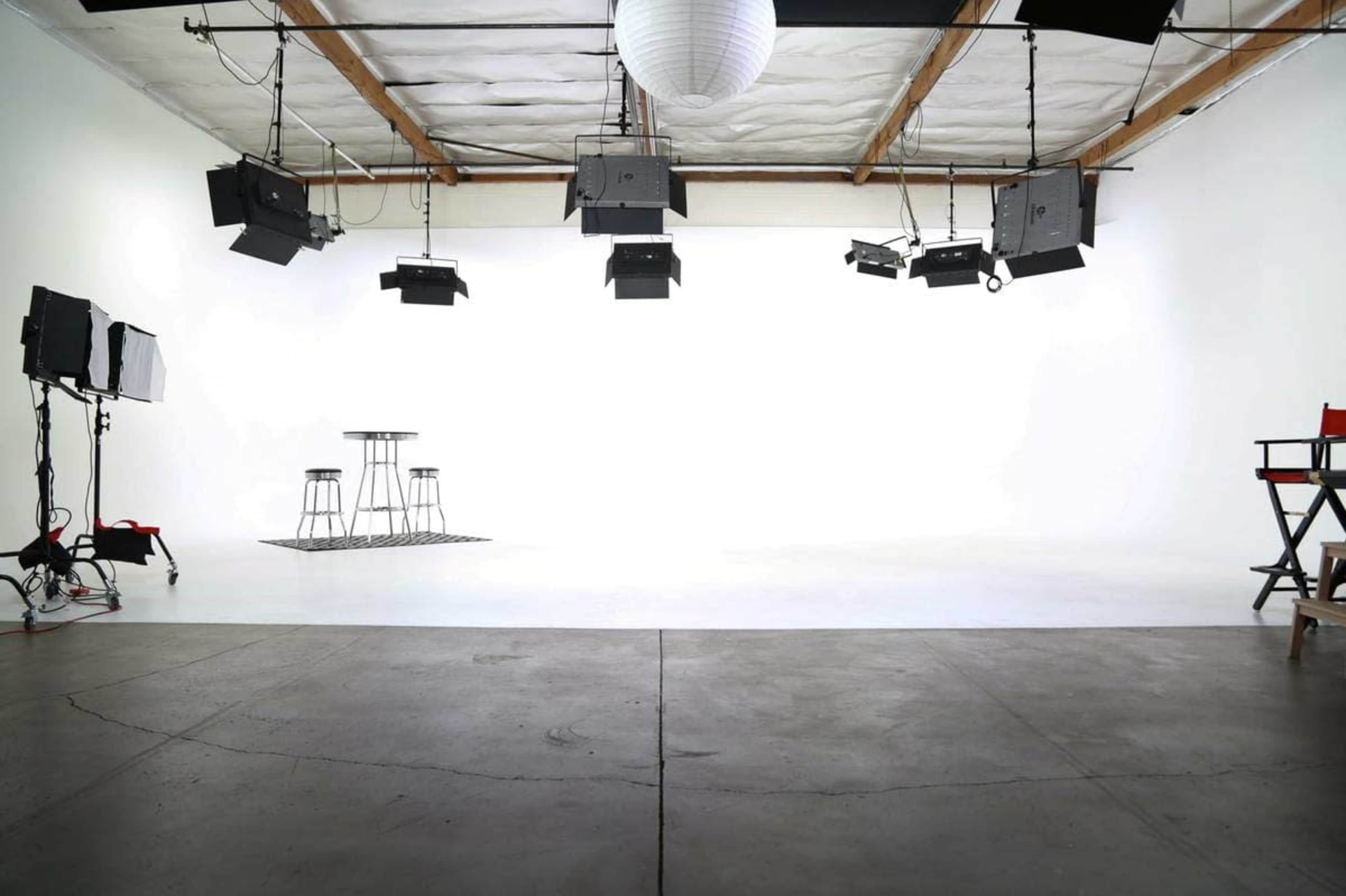 A minimally furnished studio space with two bar stools, a table, and various lighting equipment suspended from the ceiling.