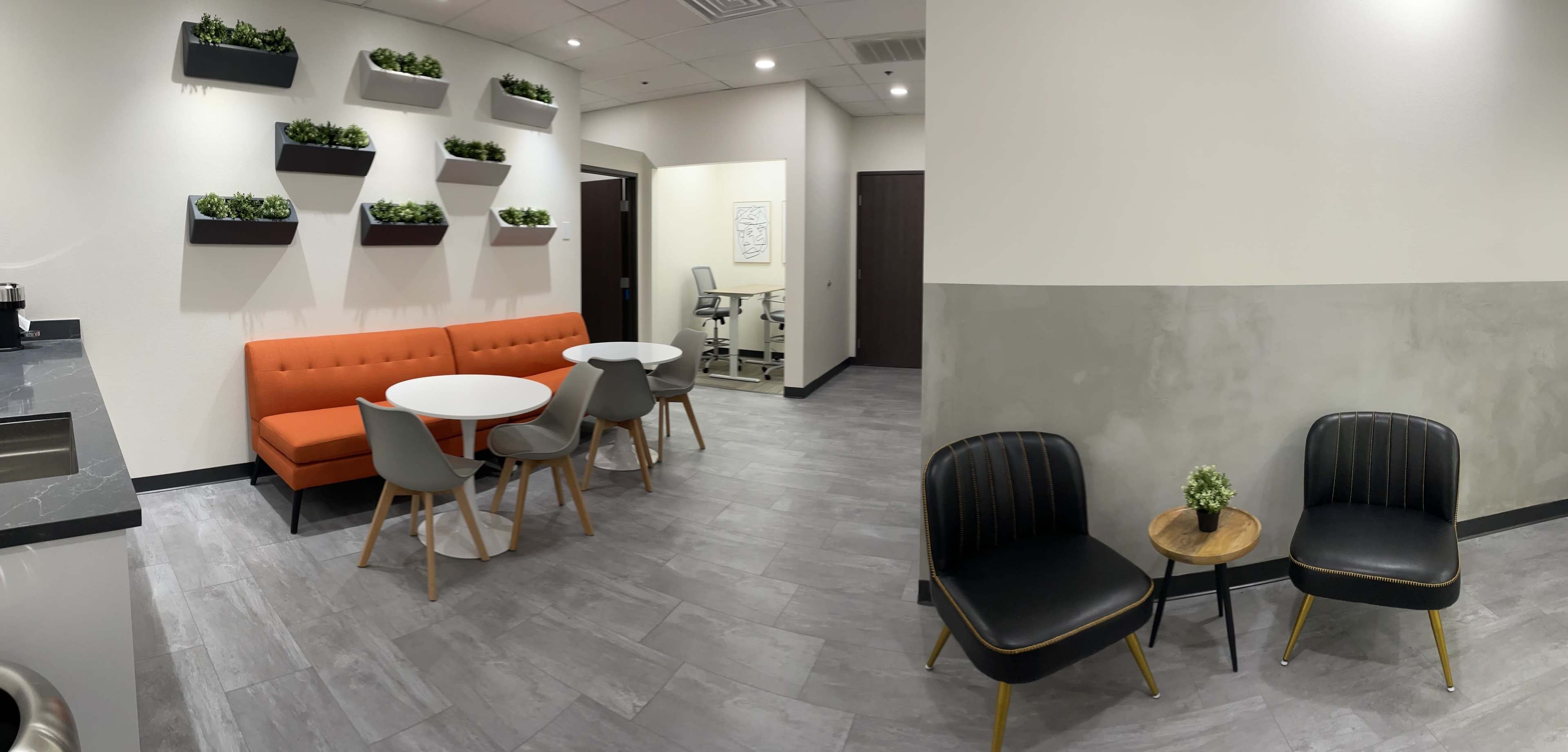 A modern waiting area featuring an orange sofa, round tables, gray chairs, and potted plants on the walls.