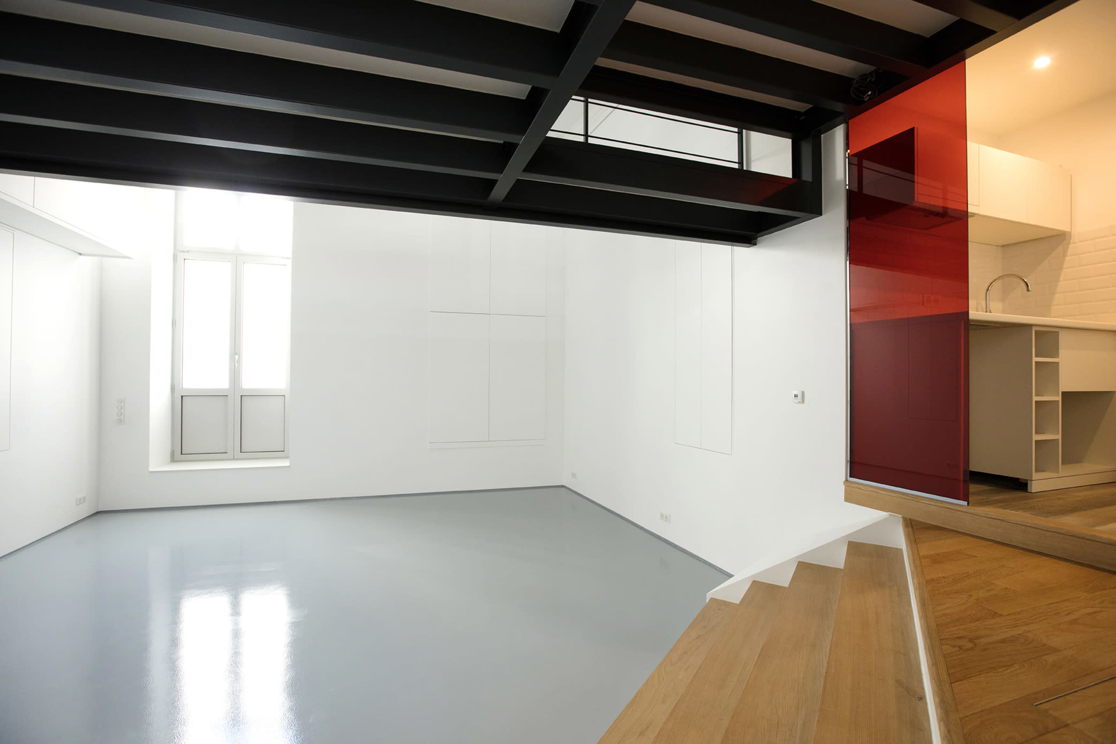 The image shows a sparsely furnished room with a polished gray floor, white walls, and a small kitchenette featuring a red accent wall.