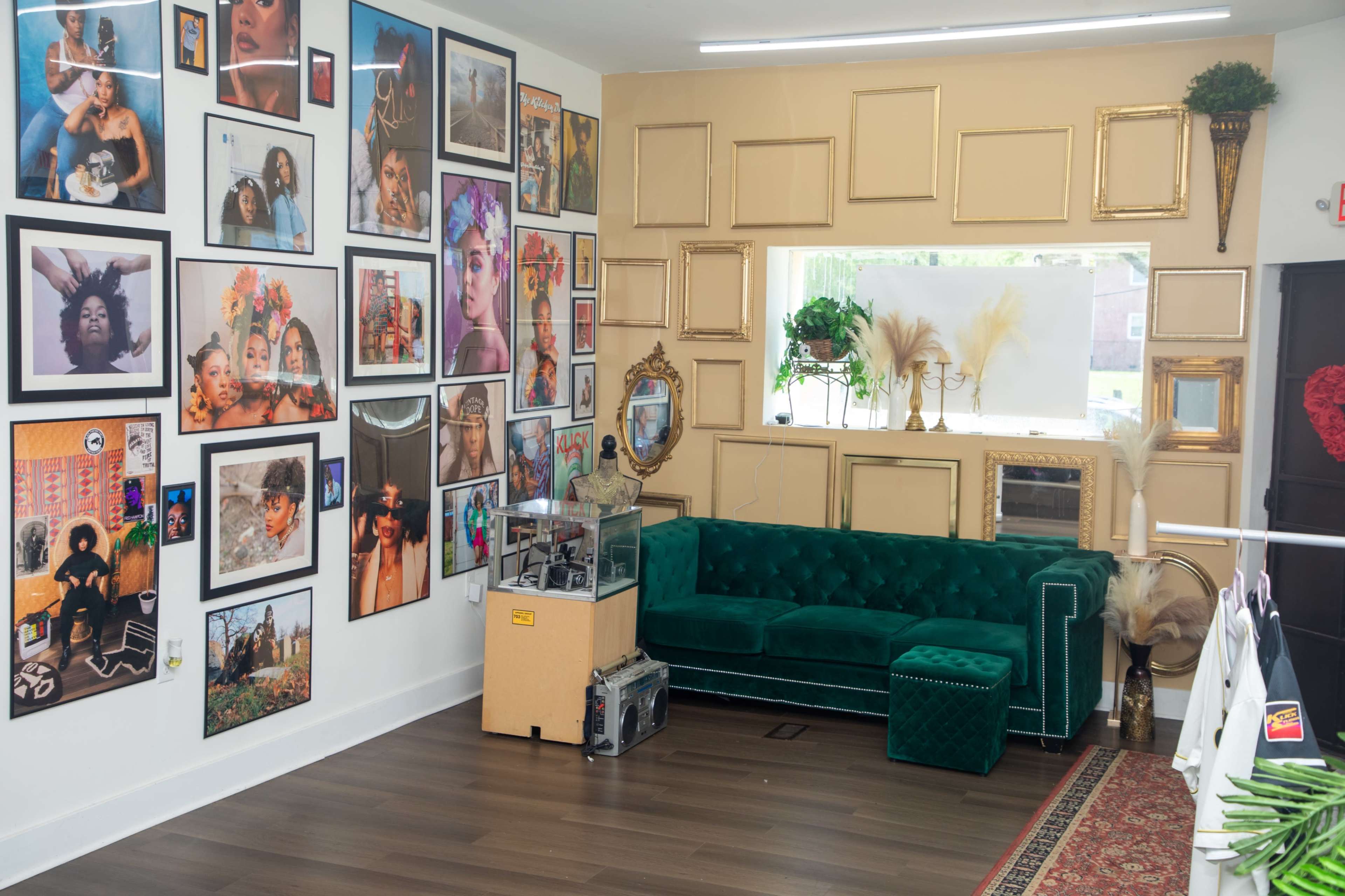 The room features a green velvet couch against a wall adorned with various framed photographs and artwork, complemented by a glass display case and decorative plants.