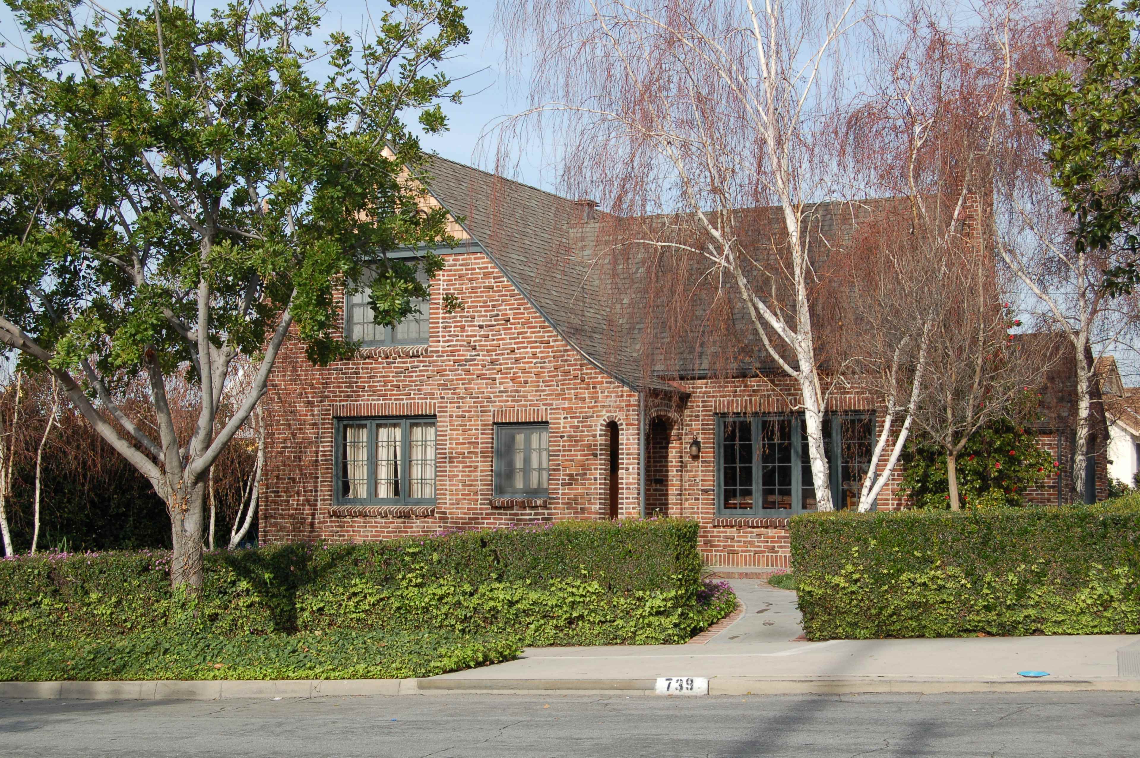 A brick house with a steep roof is surrounded by manicured shrubs and trees, located on a quiet street.